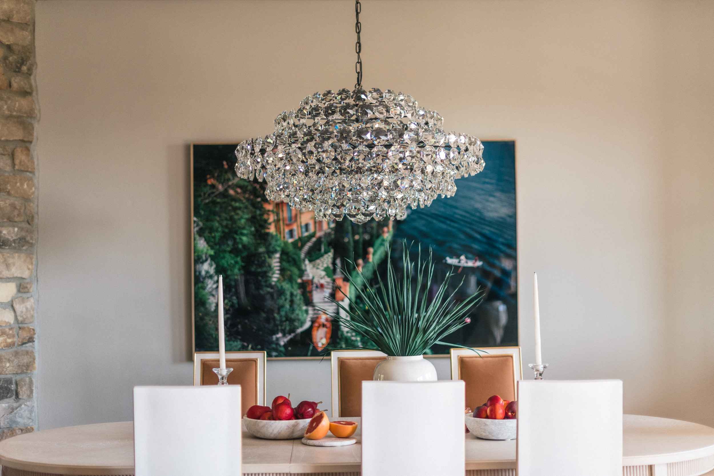Elegant dining room with a light wood table, white chairs, a large crystal chandelier, and an oversized coastal landscape painting behind the table.
