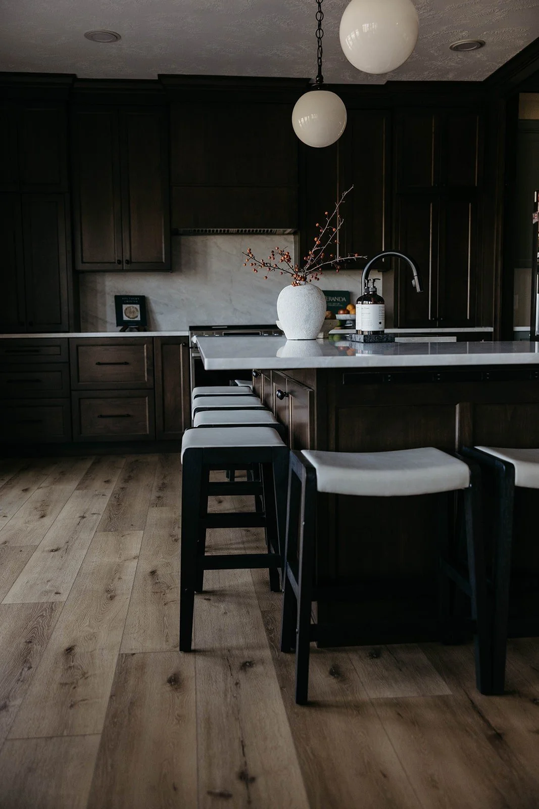 Low-angle view of the kitchen island and four dark-toned counter stools set on light, wide-plank wood flooring.