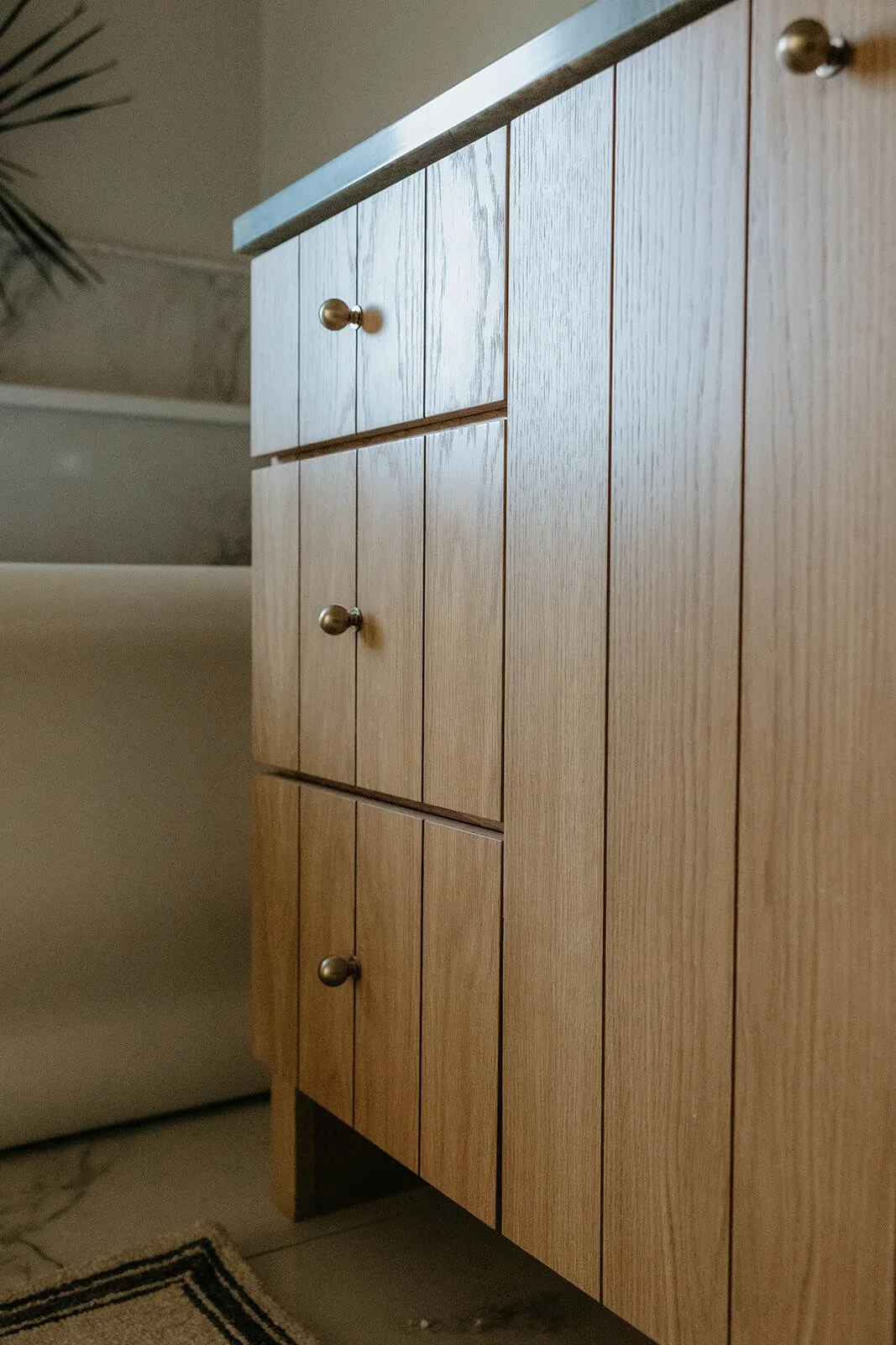 Close-up of the natural light wood vanity with vertical paneling, featuring brass knobs and a green countertop, with the freestanding tub blurred in the background.