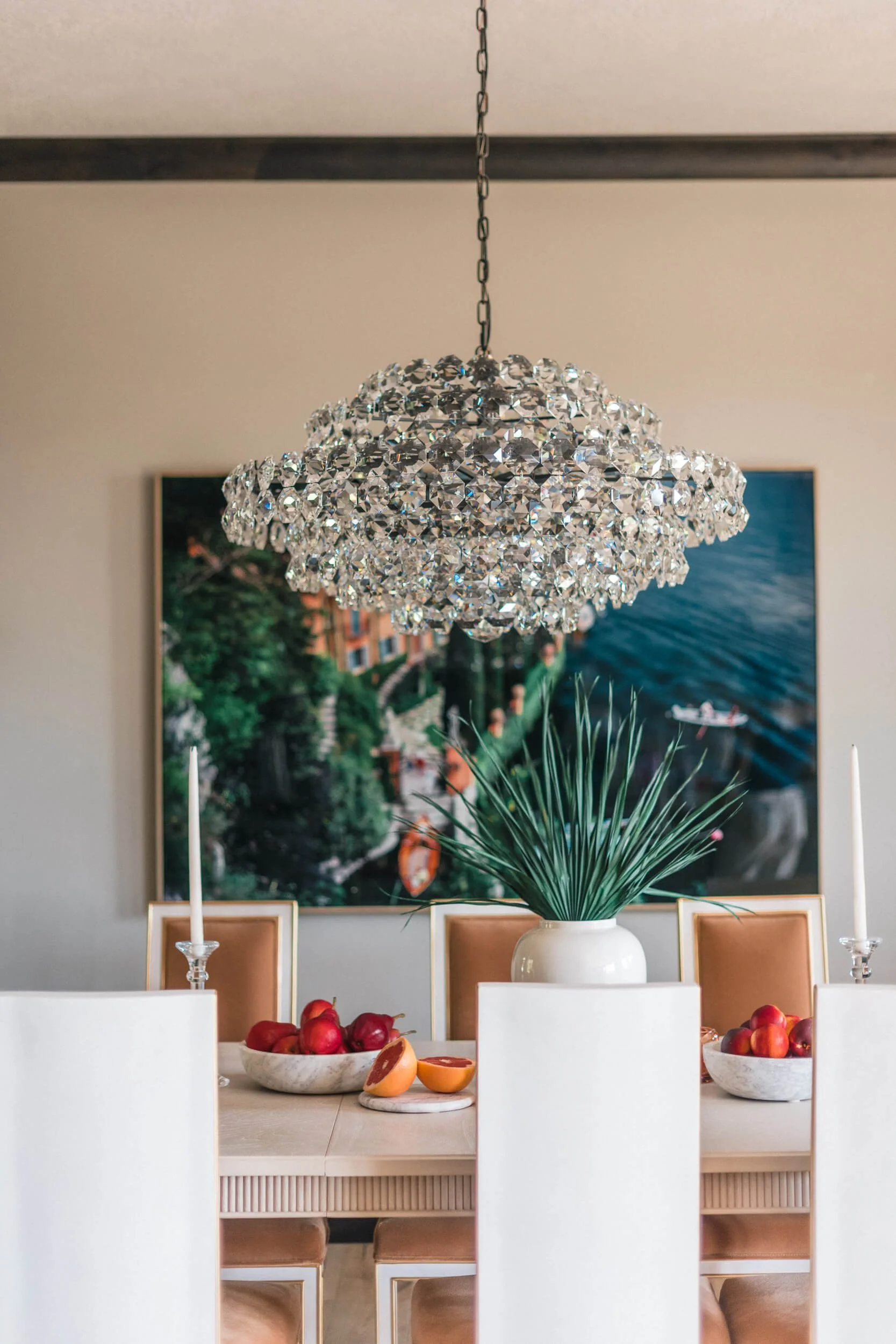 Bright, modern dining space featuring a striking crystal chandelier centered over a light wood table set with fruit and candles, with a large coastal artwork on the wall.