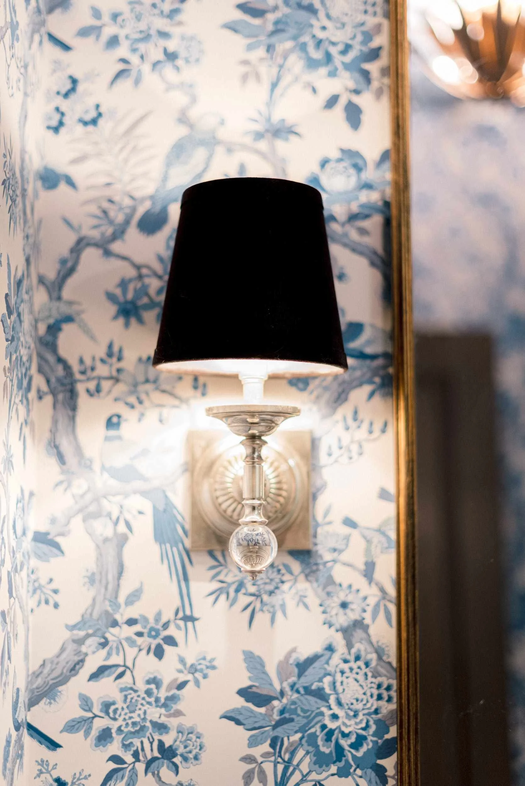 Close-up of a black-shaded wall sconce with a crystal base, set against the detailed blue and white toile wallpaper
