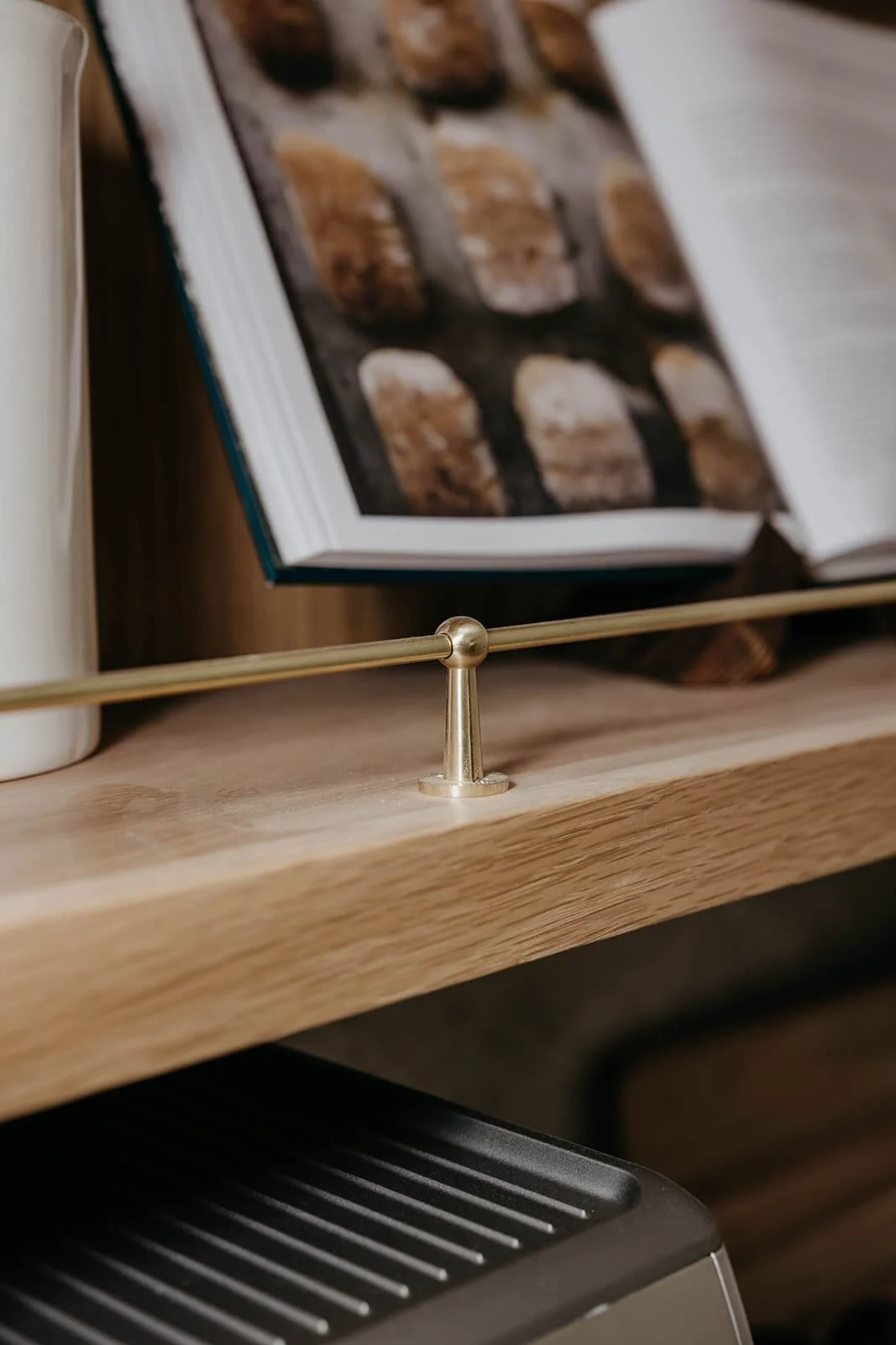 Close-up of a cookbook opened to a recipe, propped up on a light wood shelf with a brass rail above a coffee machine.
