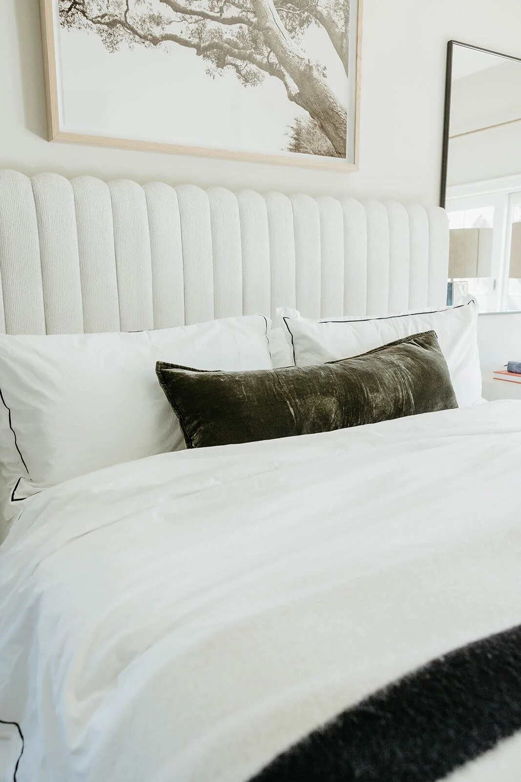 Close-up of the luxurious white bedding and channel-tufted headboard, styled with a dark green velvet lumbar pillow and a framed black and white tree print above.