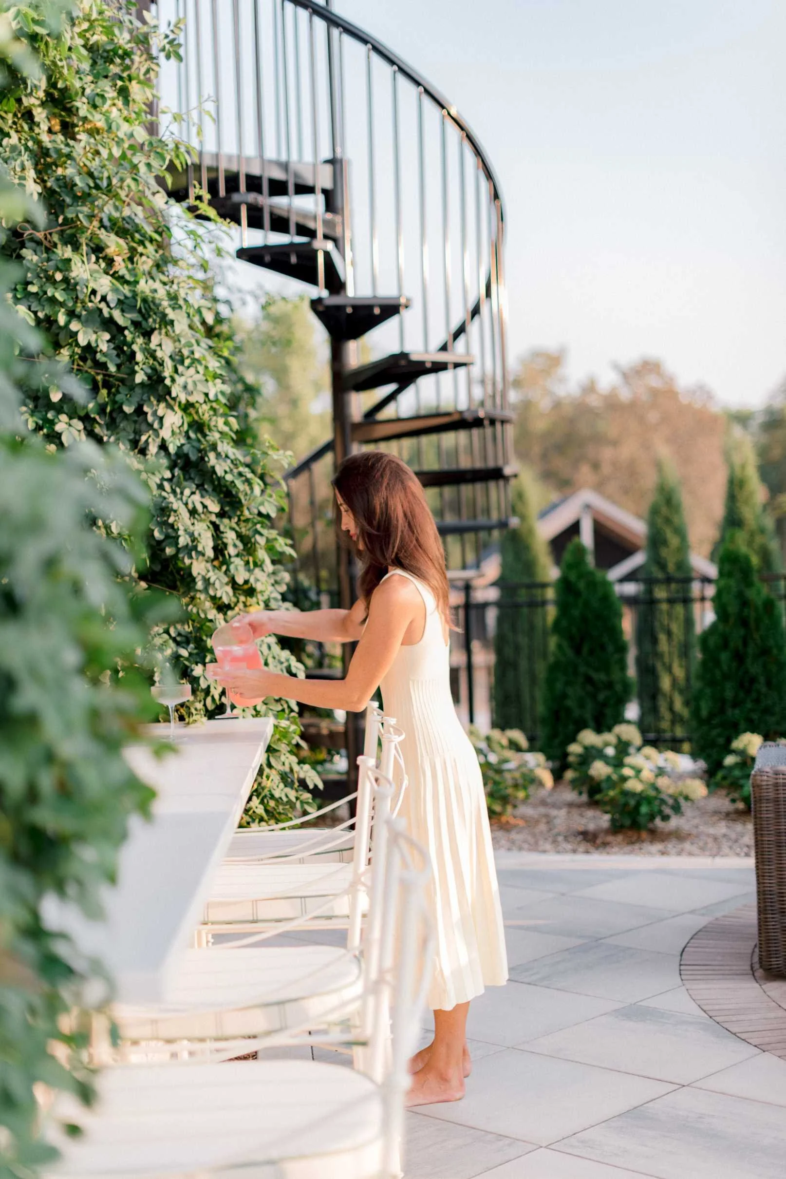 Christy Steen in a light dress serving drinks from the outdoor bar area, with a black metal spiral staircase and lush greenery visible in the background.