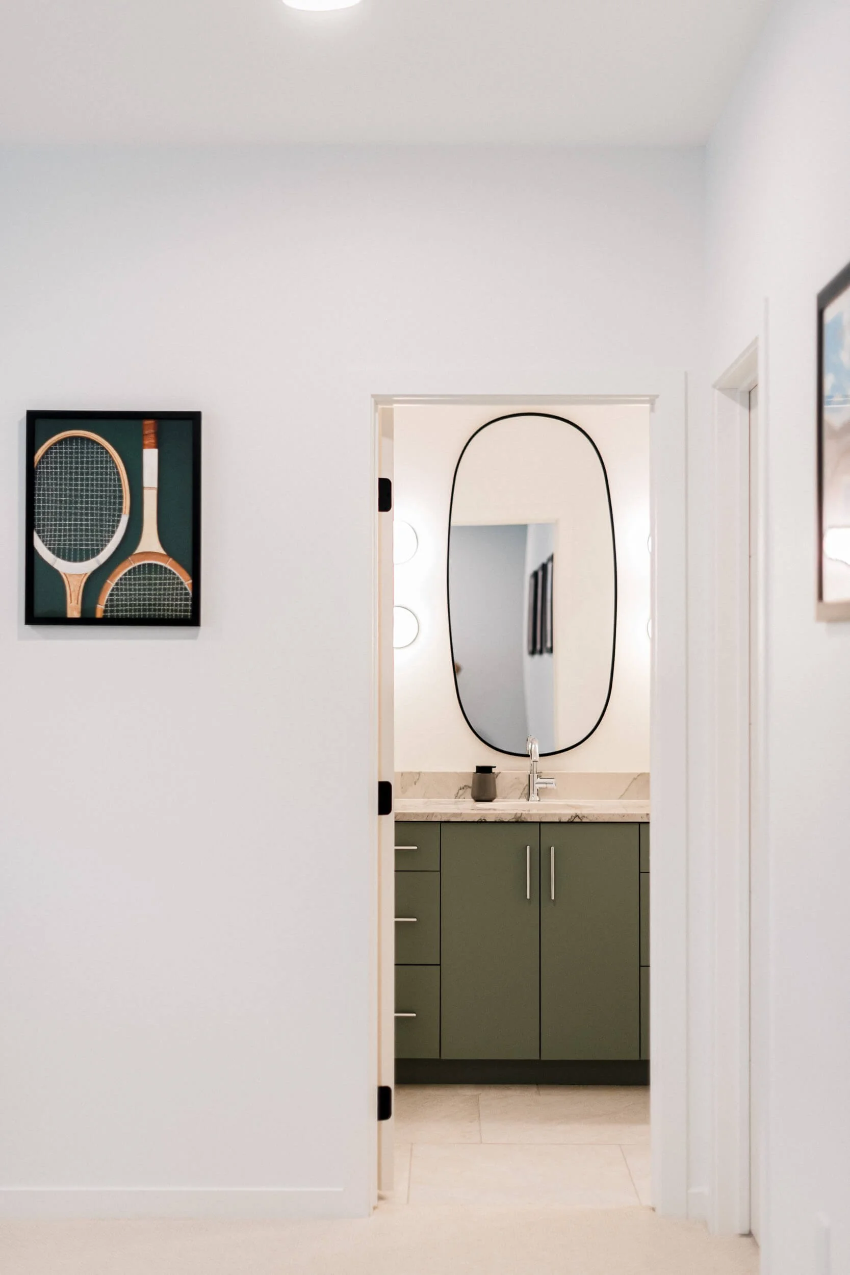 View of a hallway leading into a modern powder room with an olive-green vanity, an oval mirror, and a framed tennis racket art piece.