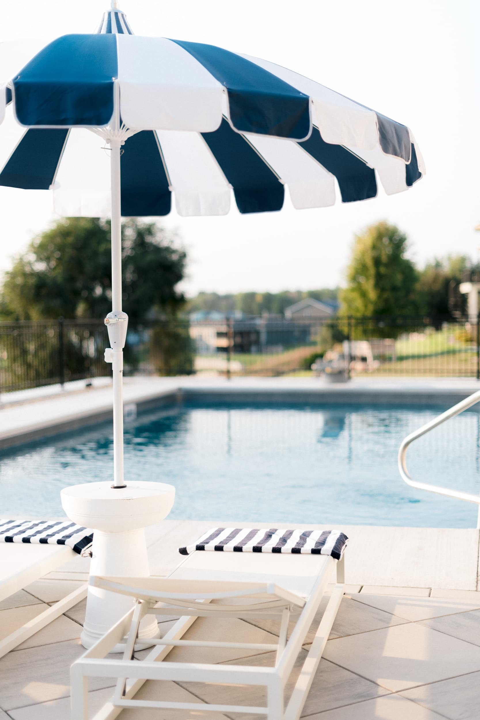 Close-up of a white chaise lounge chair with a navy and white striped cushion, under a matching striped umbrella. The pool deck overlooks a lake or large pond in the background.