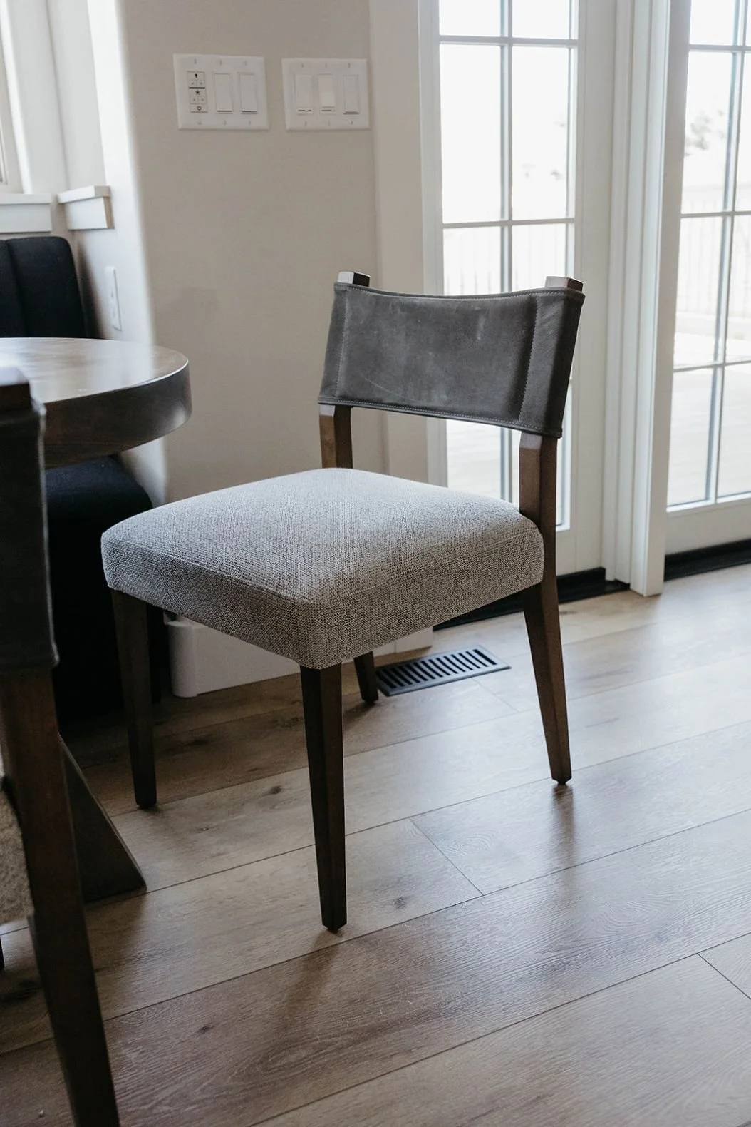 Upholstered dining chair close-up showing a gray fabric seat and a dark wood frame on light wood flooring, positioned near glass doors.