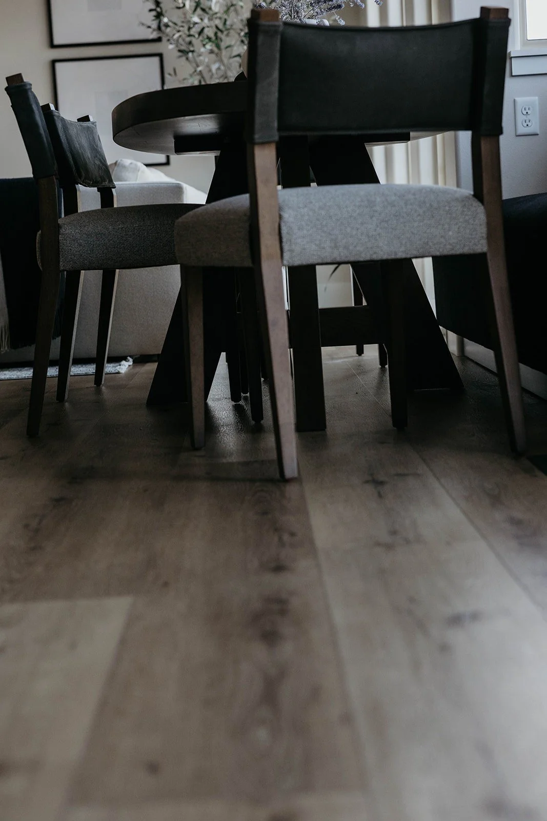 Low-angle close-up of a modern dining chair with a light gray seat and a dark wood frame, showing the legs and light wood flooring.