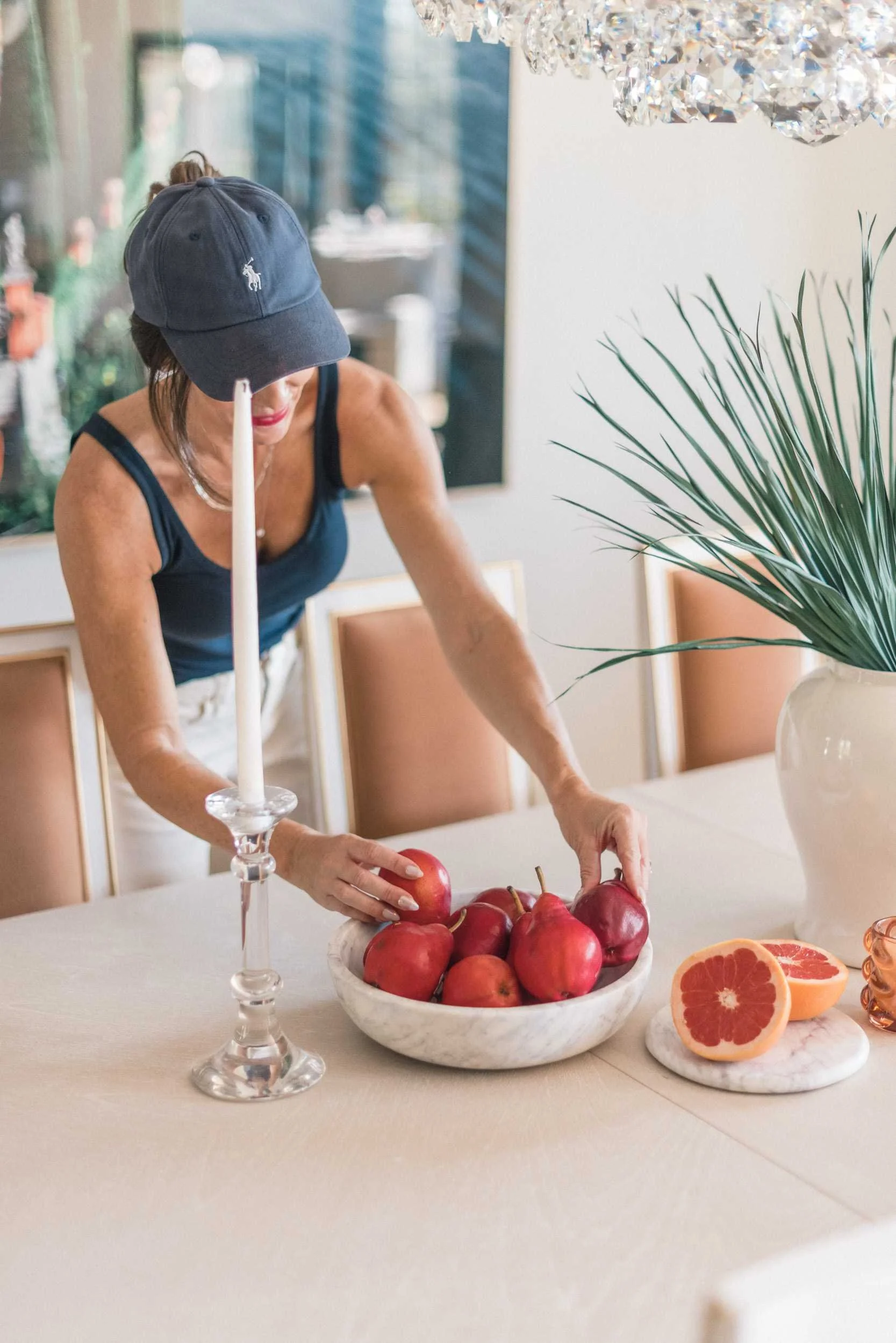 Image of a woman in a navy baseball cap styling the dining table, placing red apples into a decorative bowl.