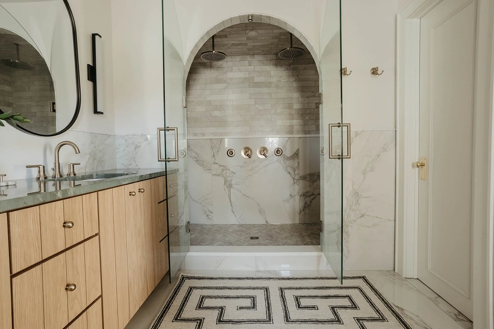 Close-up of the walk-in shower with a glass door, a dramatic arched entrance, and white marble tiling with a brass shower system.