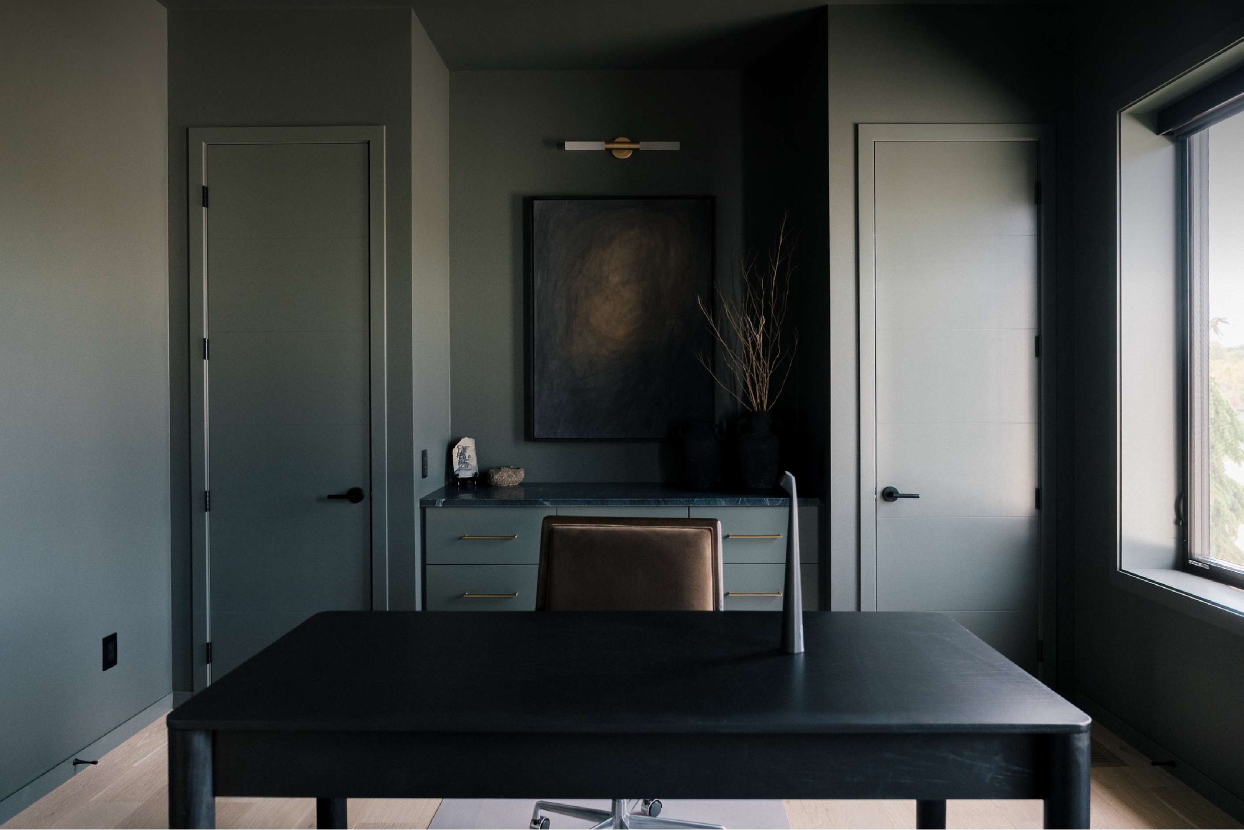 Modern office with dark green walls, a black desk, a brown office chair, a black vase with dry branches, abstract wall art, and natural light from a large window.