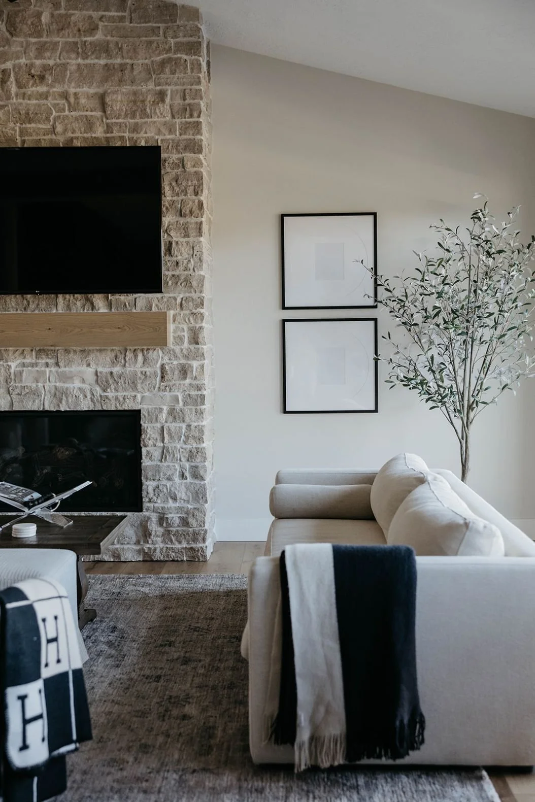 View of the light stone fireplace with a mounted TV and a wood mantel. A light sofa is styled with black and blue throw blankets and pillows.