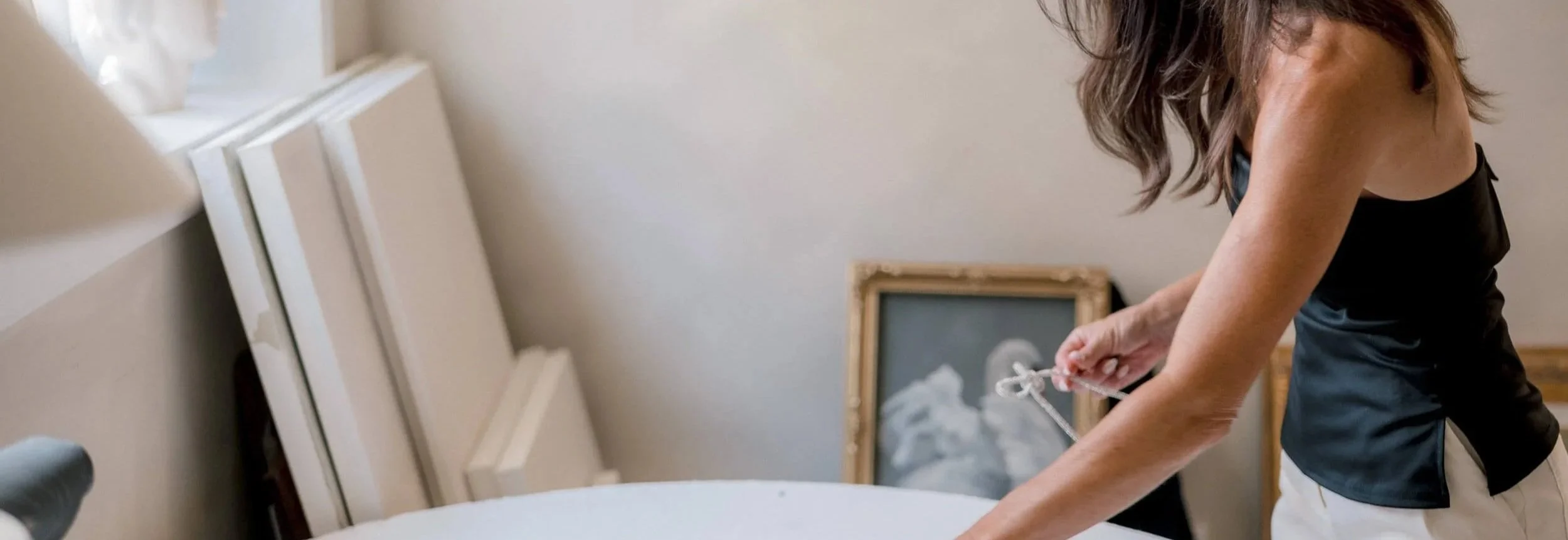 Christy Steen arranging framed paintings or photographs in a room with light-colored walls. There are additional unframed canvases leaning against the wall.