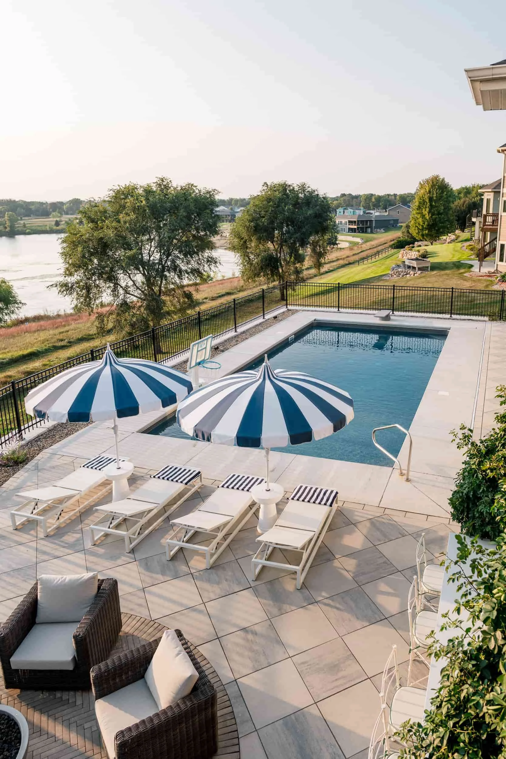 Expansive pool deck view showing white lounge chairs and gray woven outdoor armchairs. The patio overlooks a grassy hill leading down to a lake or large pond.