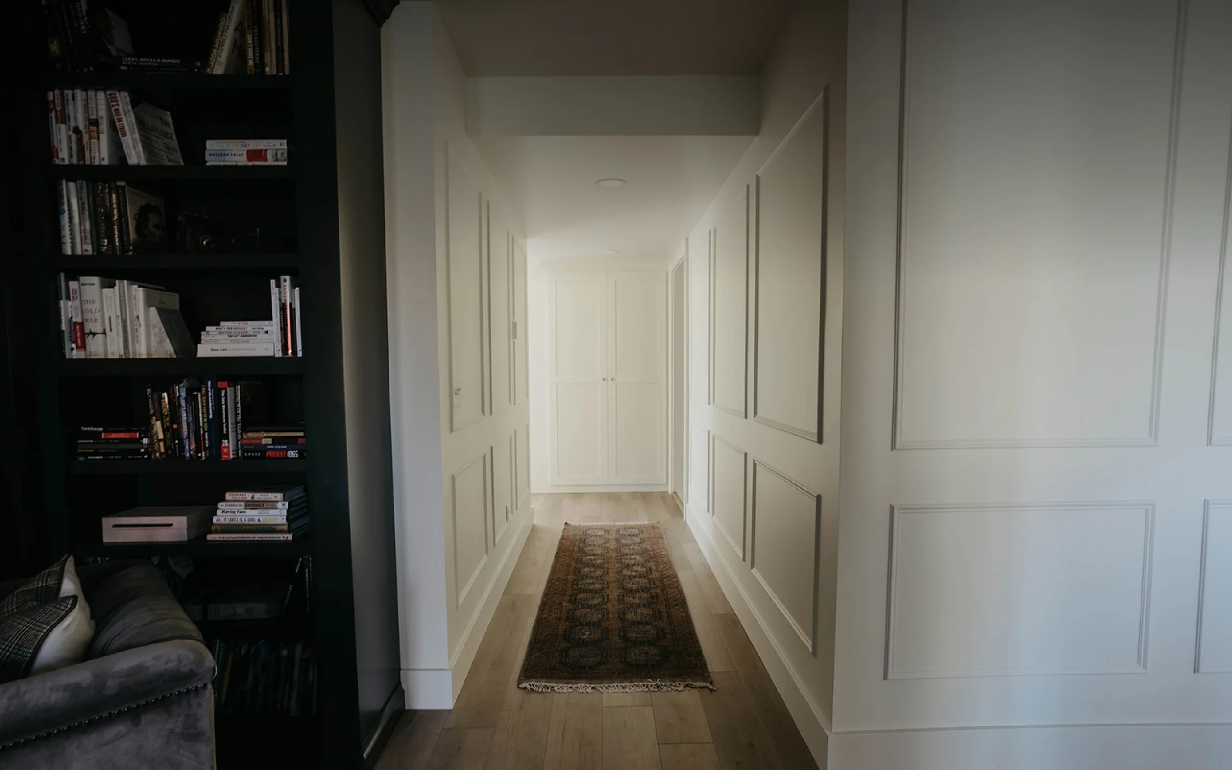 A hallway with white paneled walls, hardwood floors, a small patterned rug, white cabinets at the end, and a dark bookshelf with books on the left side.