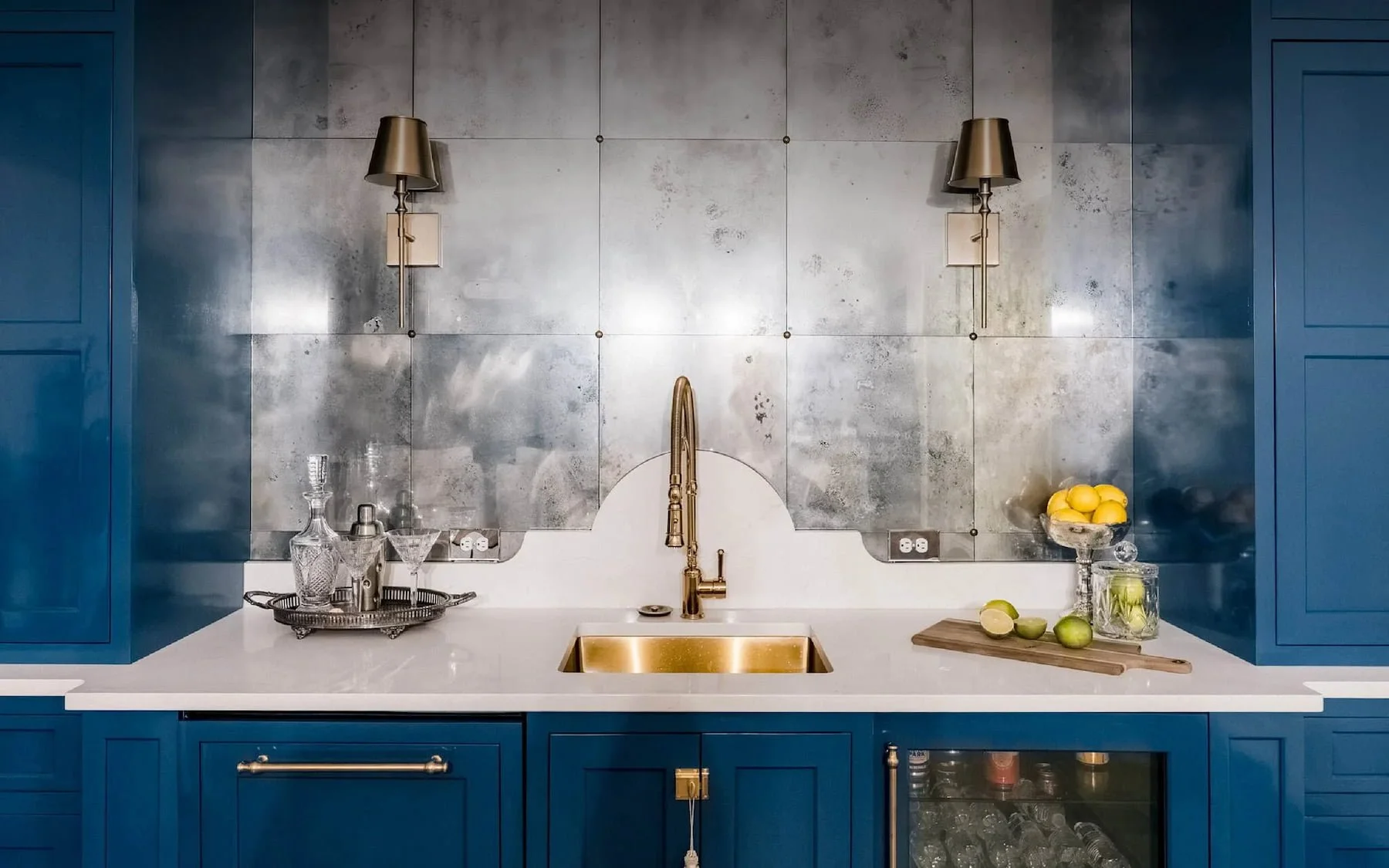 Basement bar with blue cabinets, white countertop, brass fixtures, silver tiled backsplash, and a small wine fridge.