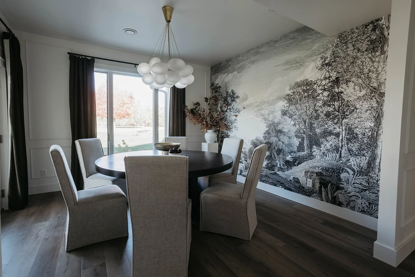 Dramatic dining room featuring a round dark wood table, gray upholstered chairs, and a striking black and white nature mural wallpaper accent wall."