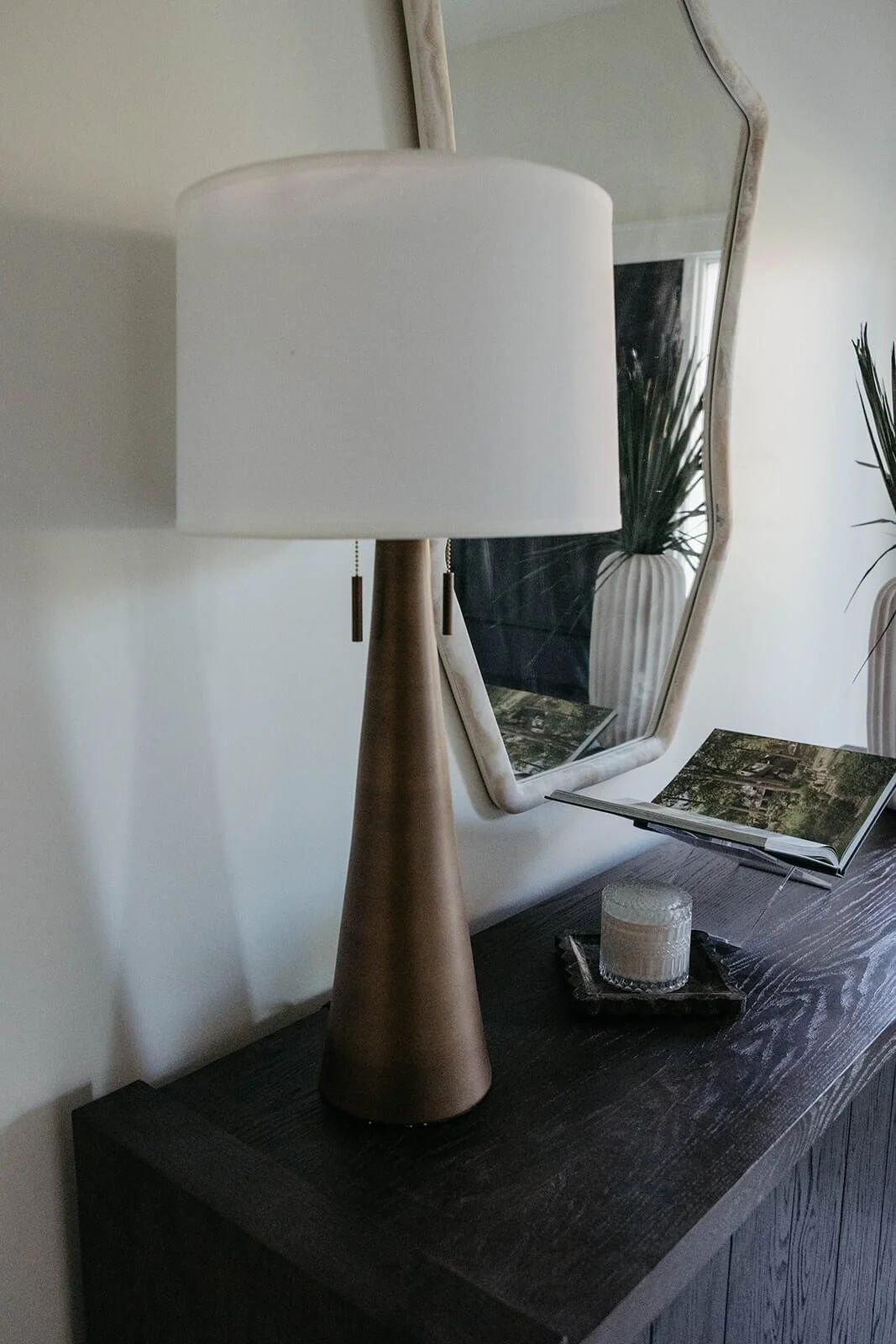 Close-up of entryway console styling showing a gold-base table lamp, a scented candle, and a white ceramic vase with greenery, reflected in a unique-shaped mirror.