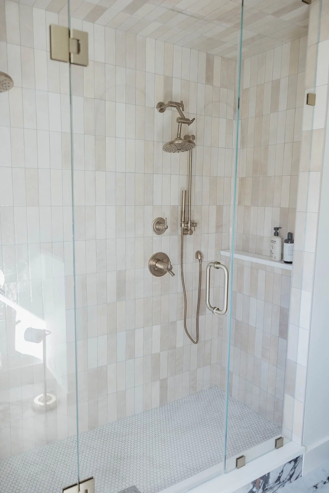 Walk-in shower with clear glass door, featuring vertical stacked white subway tile and a brushed nickel shower system. The floor is a small white hex tile.