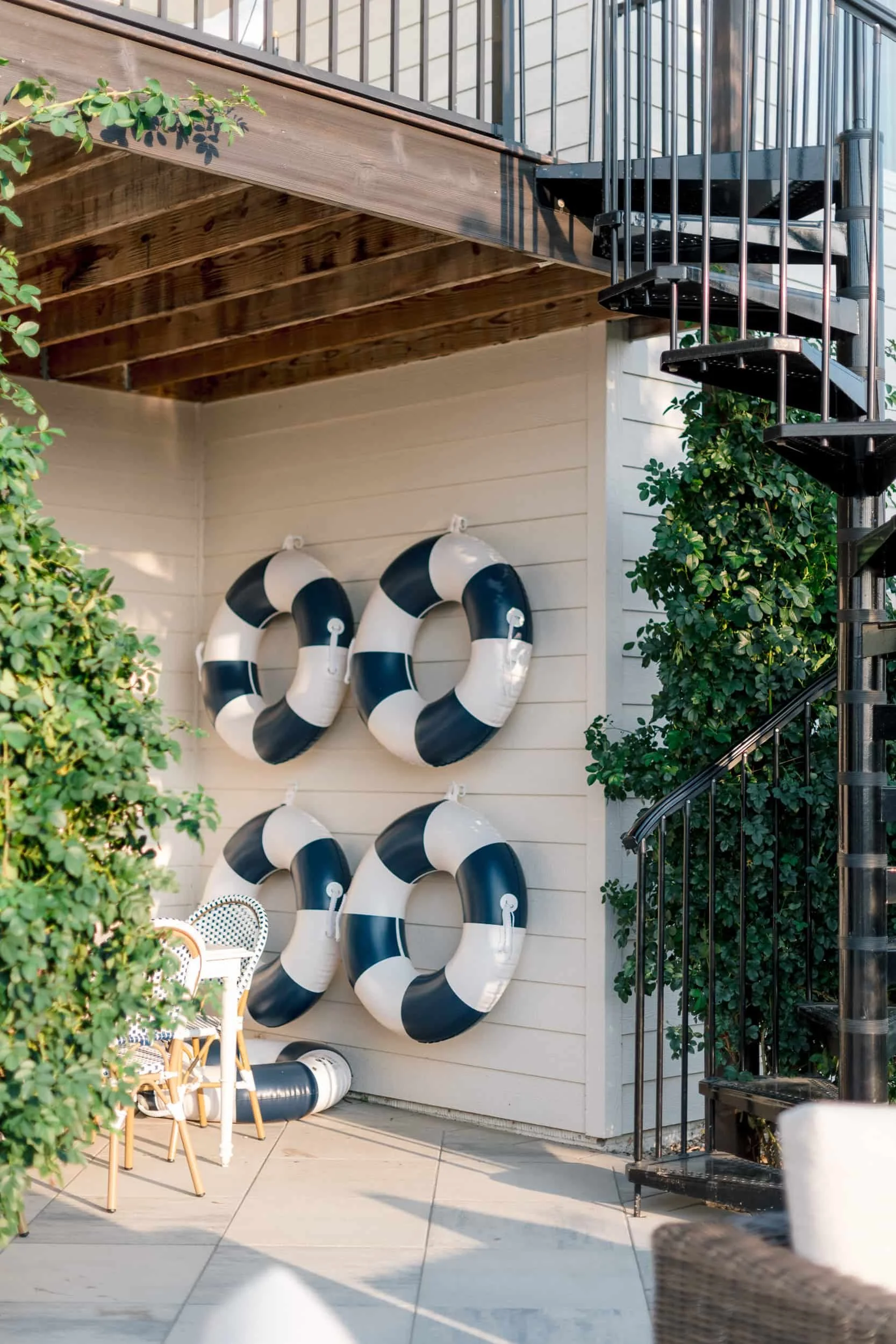 Outdoor entertaining space showing a white shiplap wall with life preserver decor, a white bar counter, and a black metal spiral staircase to the right, framed by lush climbing vines.