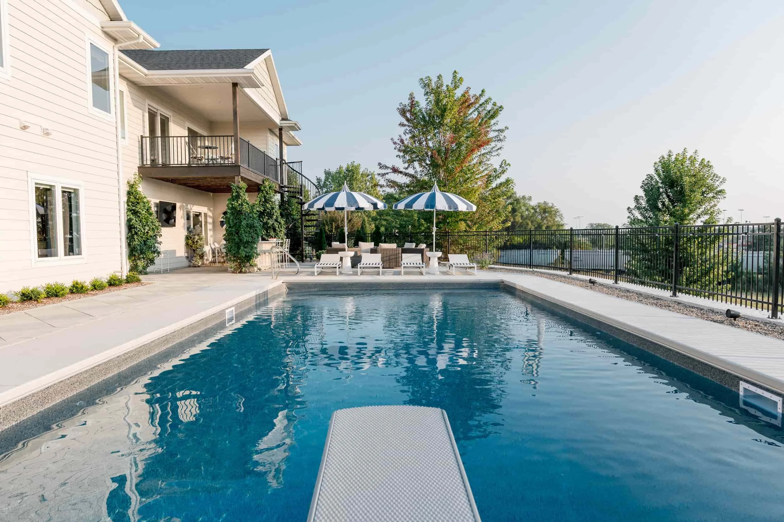 Backyard view of a modern house with a large, rectangular swimming pool that includes a diving board. The pool area is styled with striped umbrellas and is enclosed by a black metal fence.
