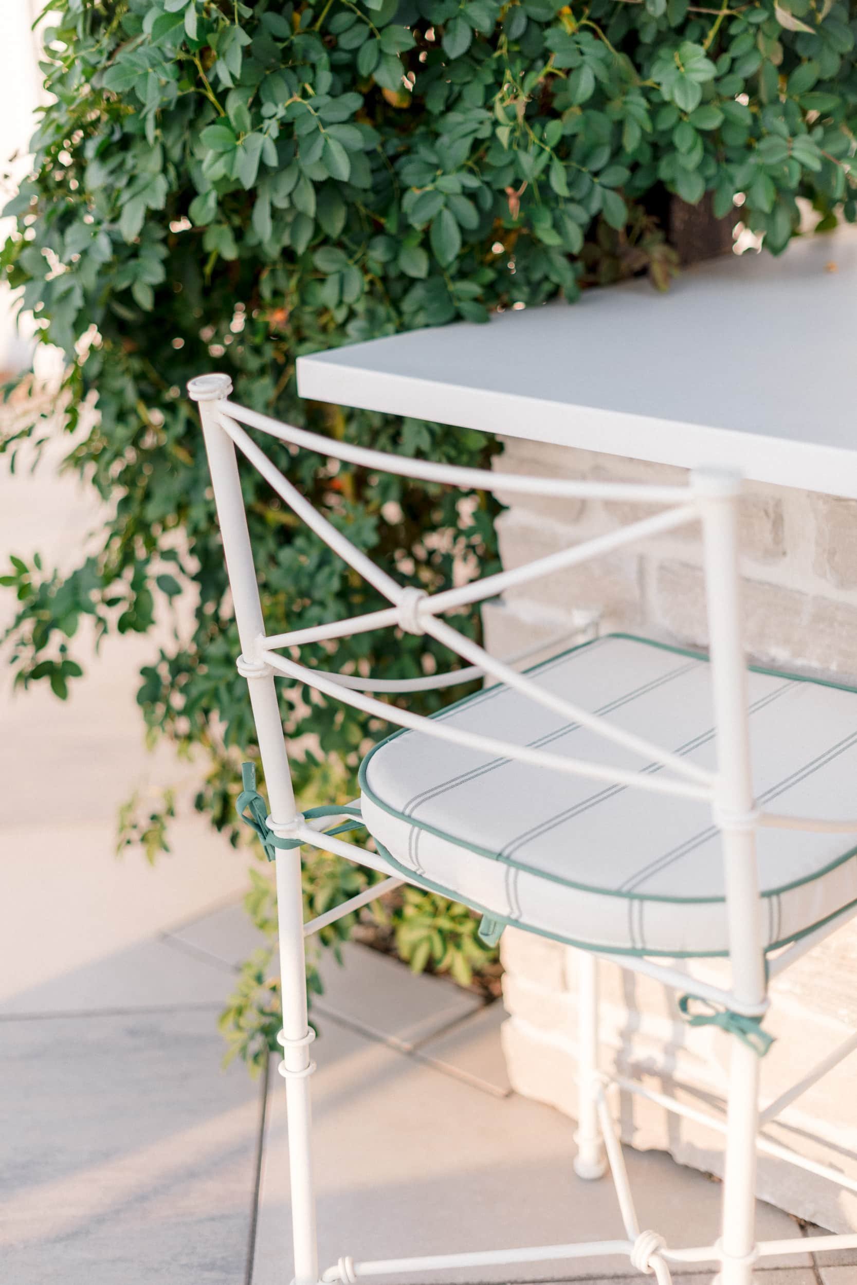 Close-up of a white metal barstool with a striped cushion at the outdoor bar, set against a stone and vine-covered wall.