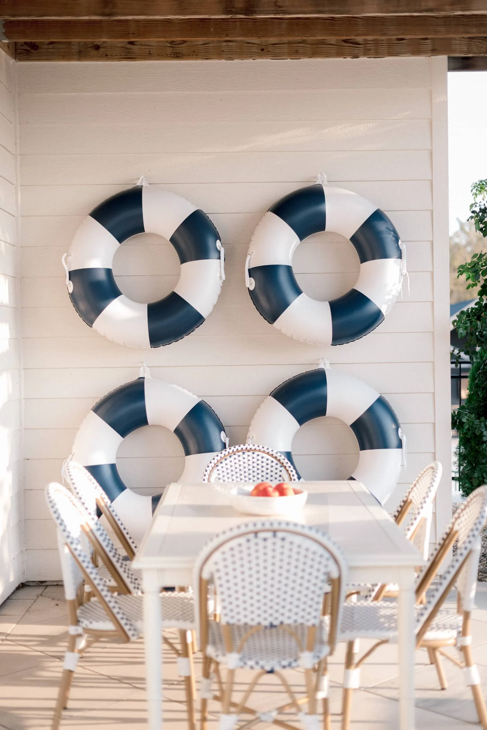 Close-up of a nautical-themed outdoor dining area. Four large navy and white life preservers are mounted on a white shiplap wall above a dining table and woven chairs.