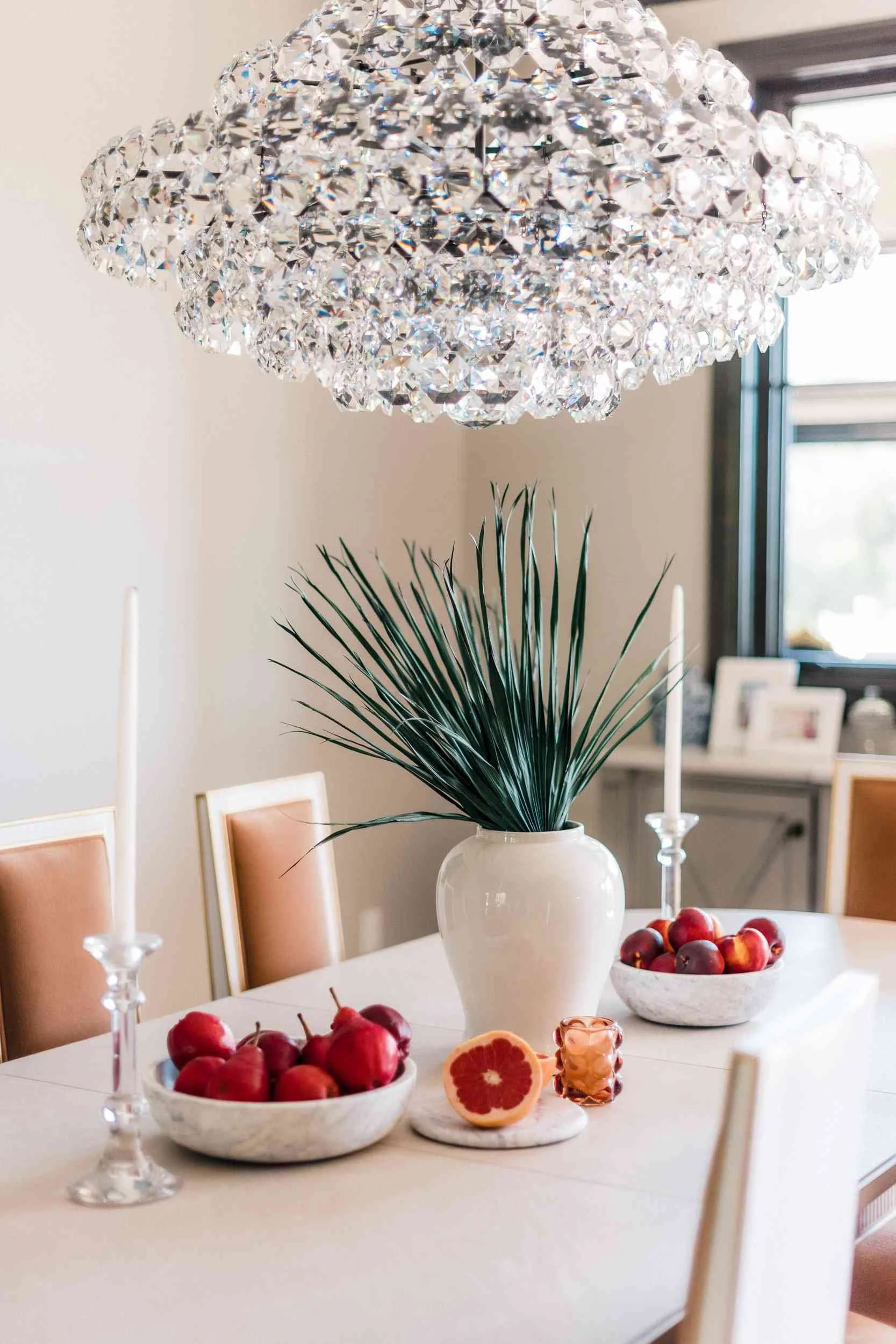 Detailed close-up of the crystal cloud chandelier hanging over the center of the dining table styled with a white vase of greenery, bowls of fruit, and tall white candles. 