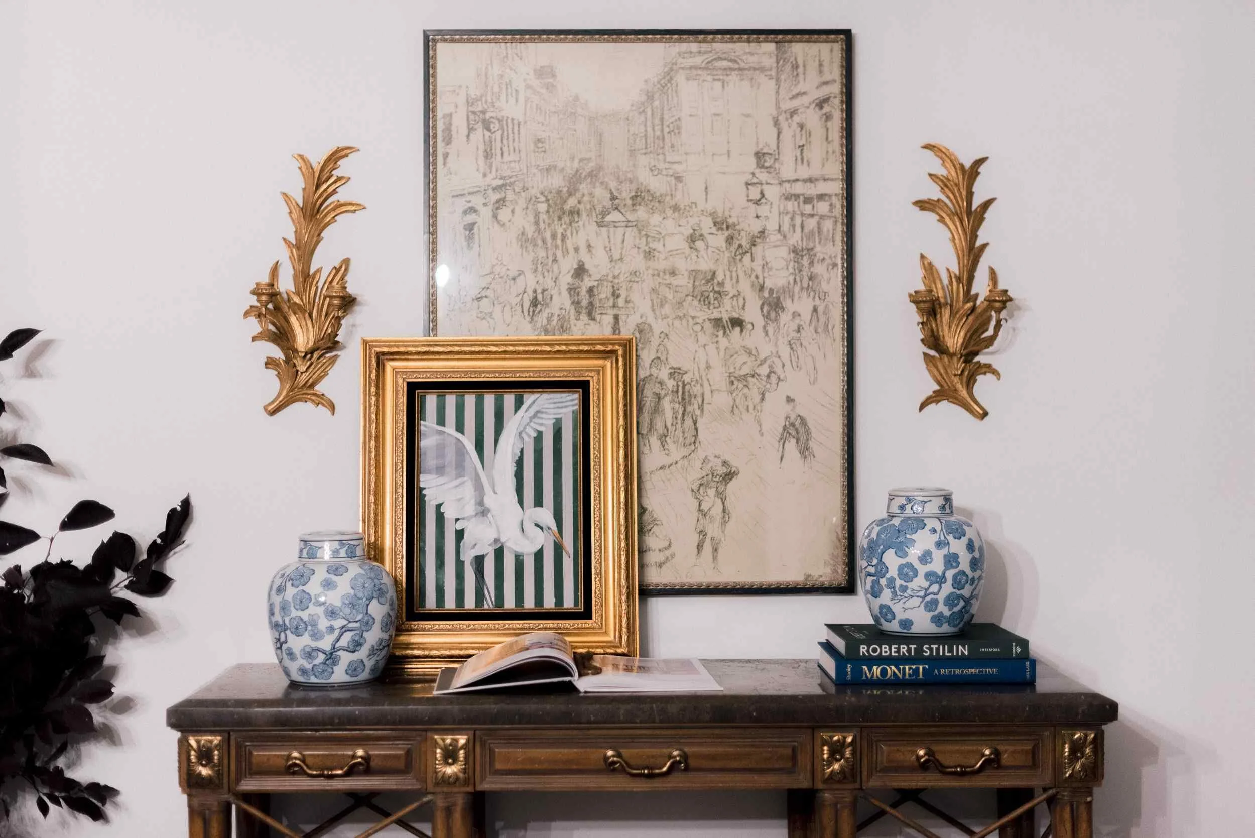 Close-up of the styled console table, showing the framed original artwork by Christy Steen, stacked blue books, and a gilt sconce on the white wall.