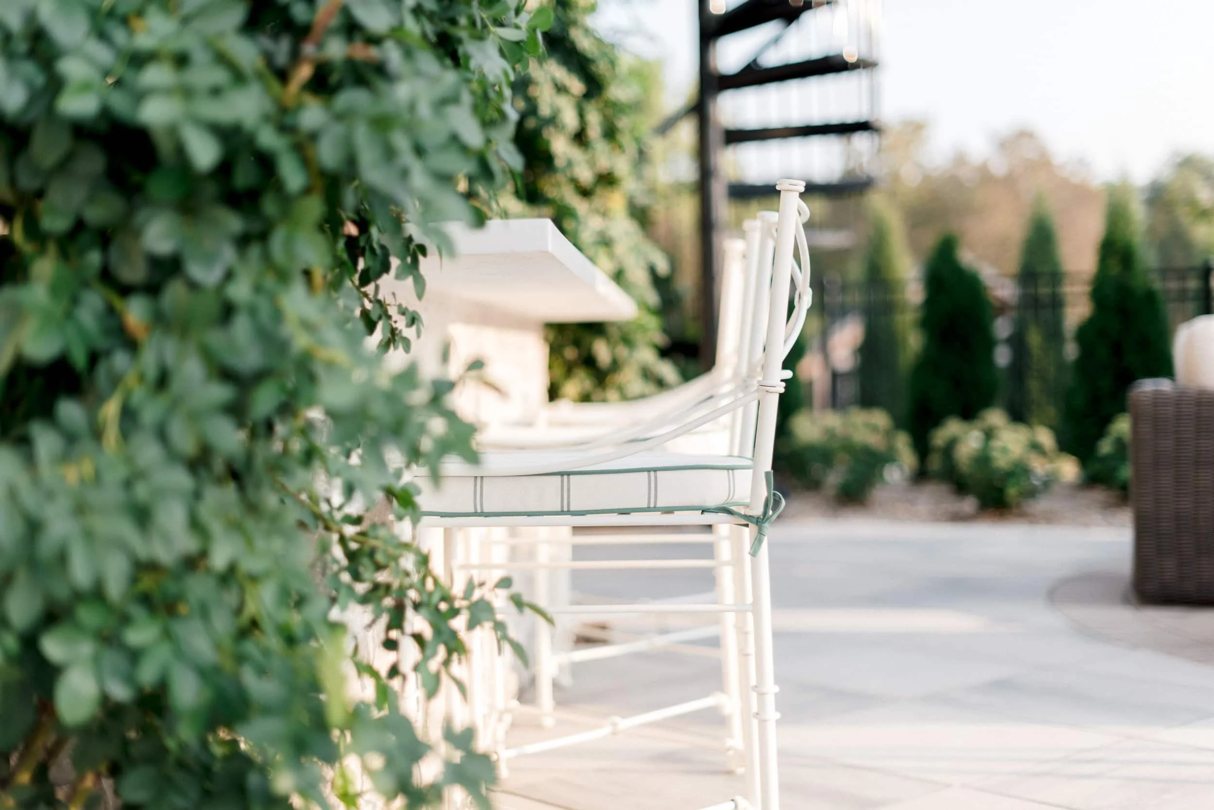White metal barstool on a patio with a distant view of the landscaped backyard and tall trees.