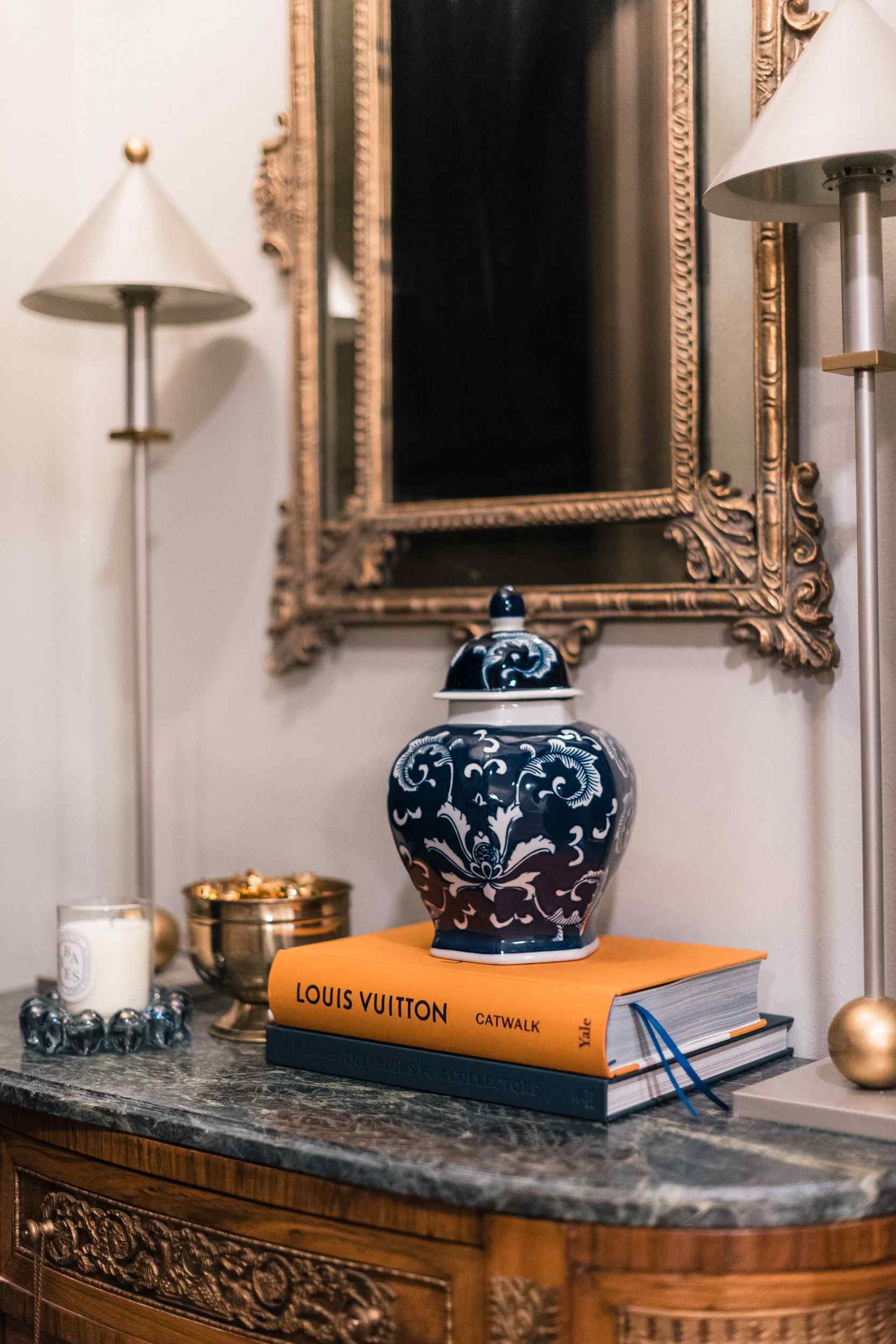 Detail of the marble-top console, showing a stack of books, a white candle, and the intricate carved wood front.