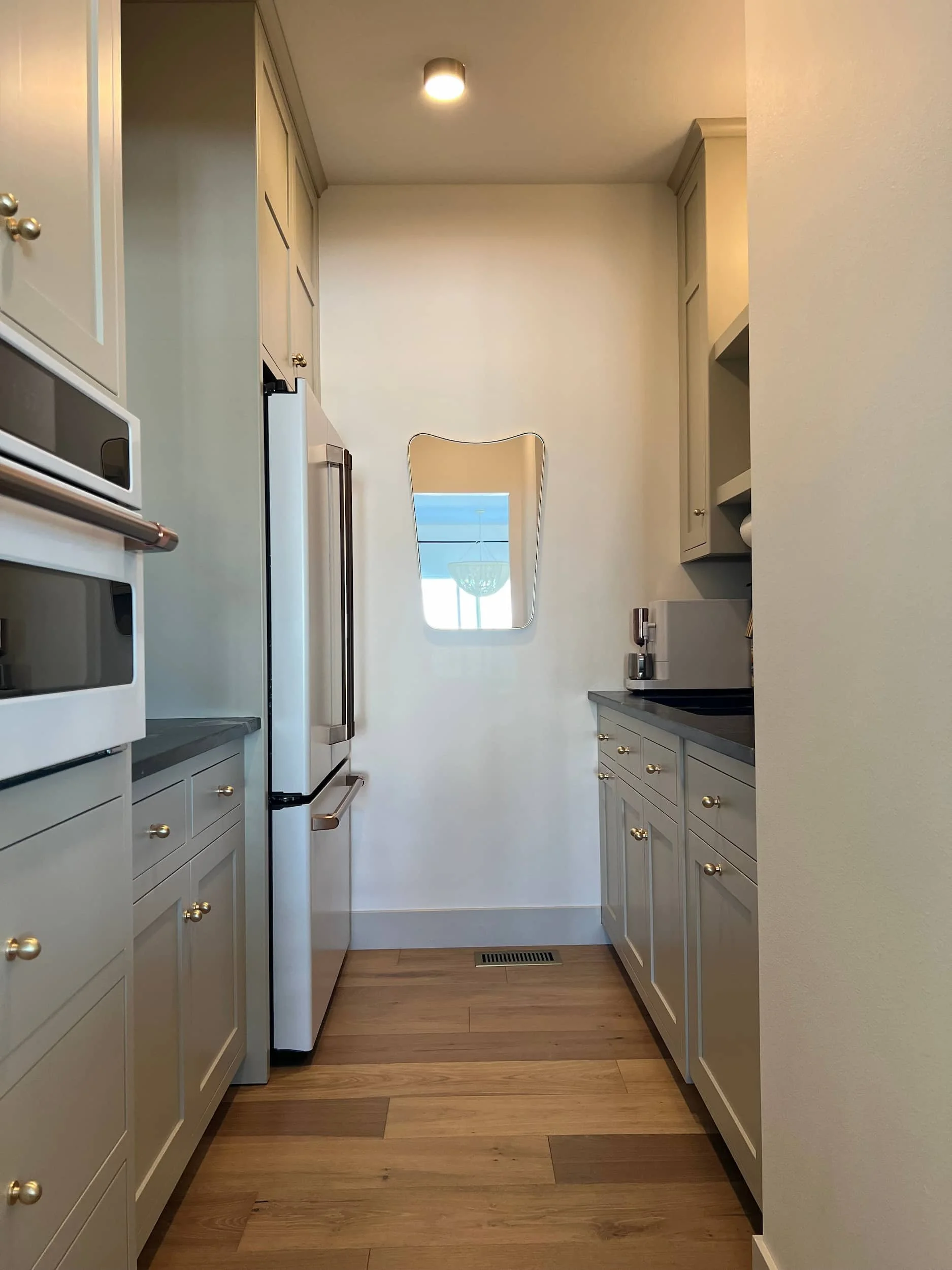 Narrow galley kitchen or hallway featuring light sage green cabinetry with brass hardware, a dark countertop, and a small decorative arched mirror on the wall.
