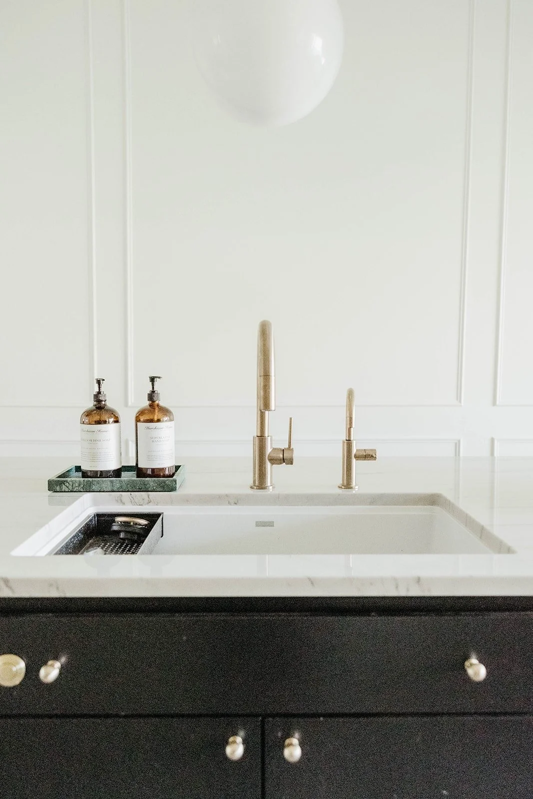 Close-up of the kitchen sink with a brushed gold faucet set against dark cabinetry and a white paneled wall. Brass knobs provide accent.
