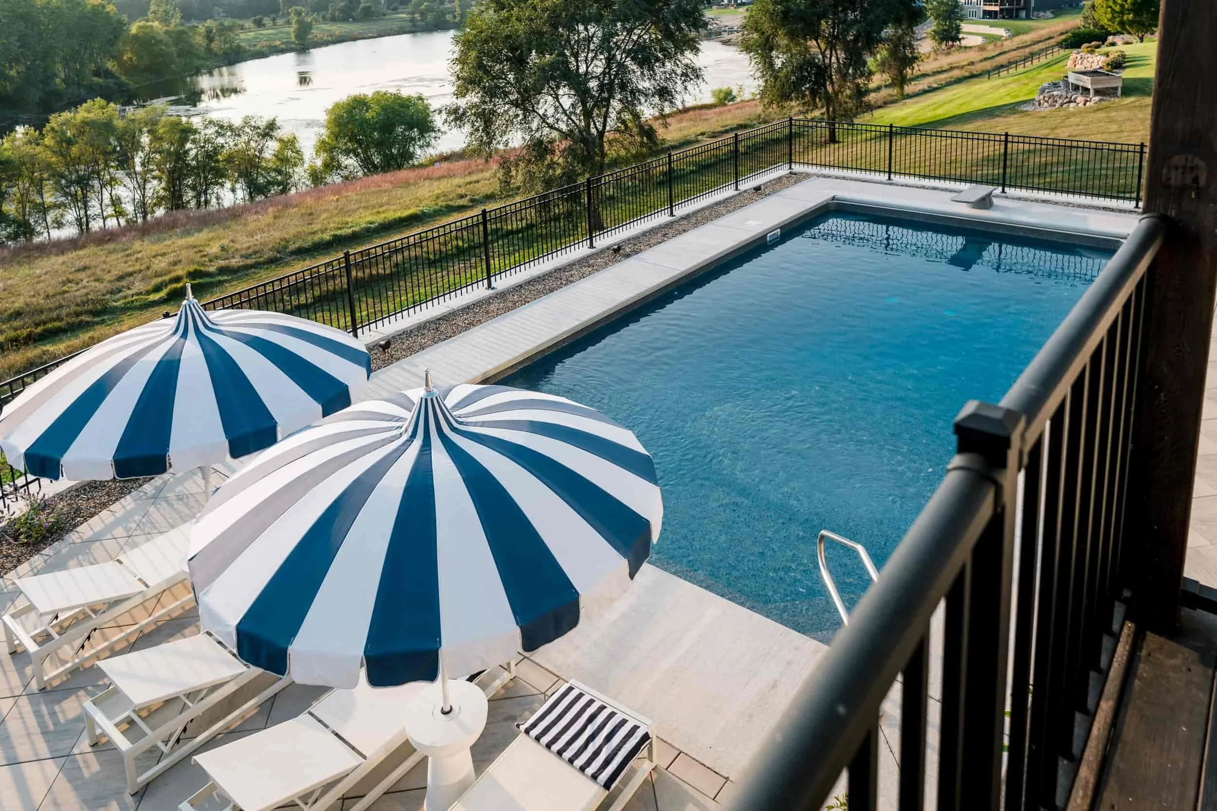 Aerial view of a rectangular in-ground swimming pool with a black metal fence, overlooking a body of water and the distant landscape. Two navy and white striped umbrellas are visible.