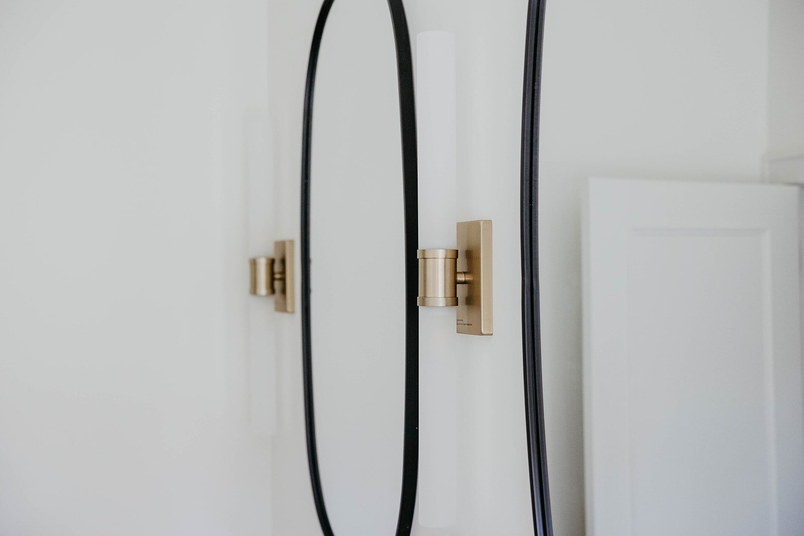 Close-up of a black oval mirror and a modern brass and glass wall sconce mounted on a white wall above the vanity.