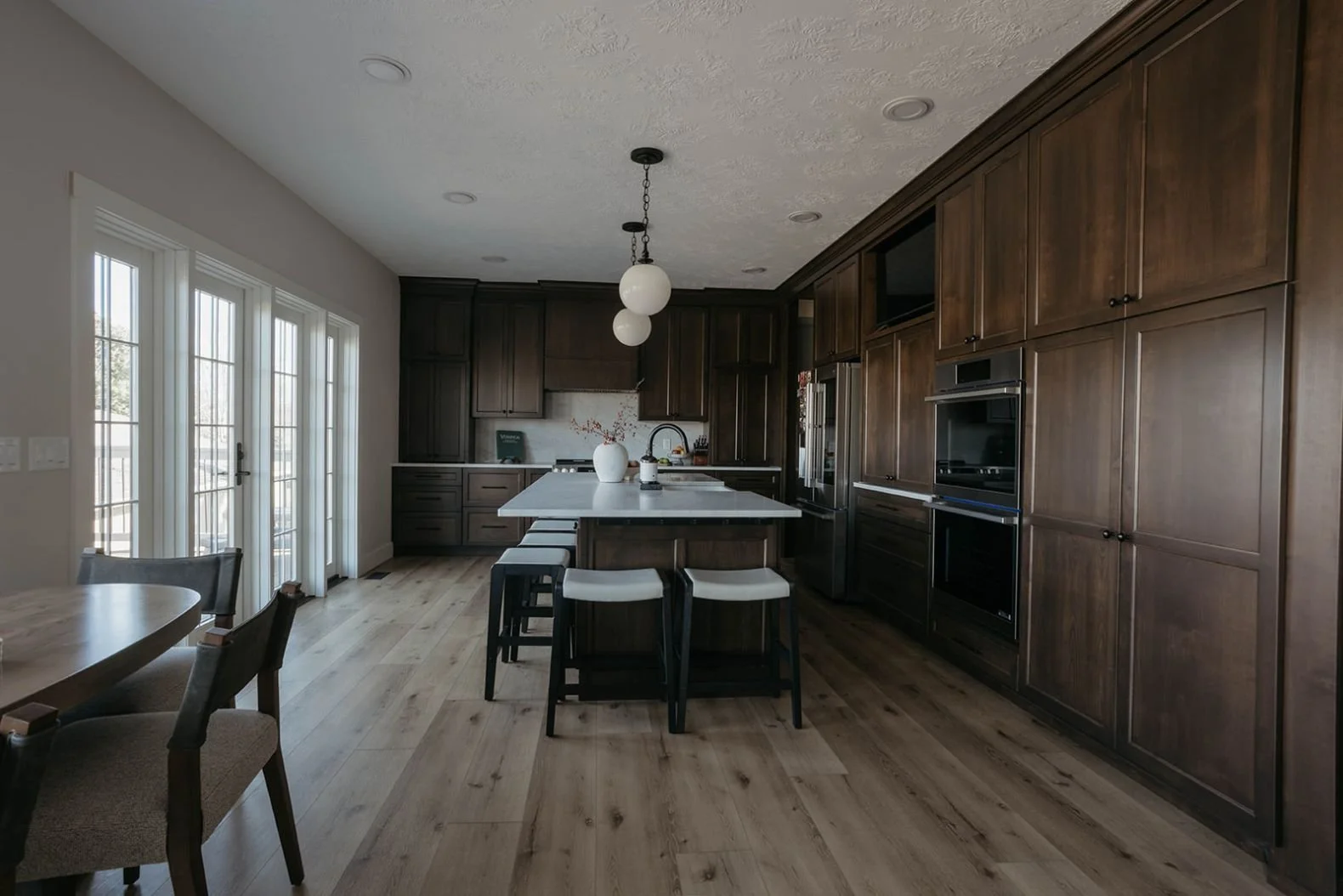 View of the dining area and dark wood kitchen with large glass doors leading outside. The dining table features a white pendant light overhead.
