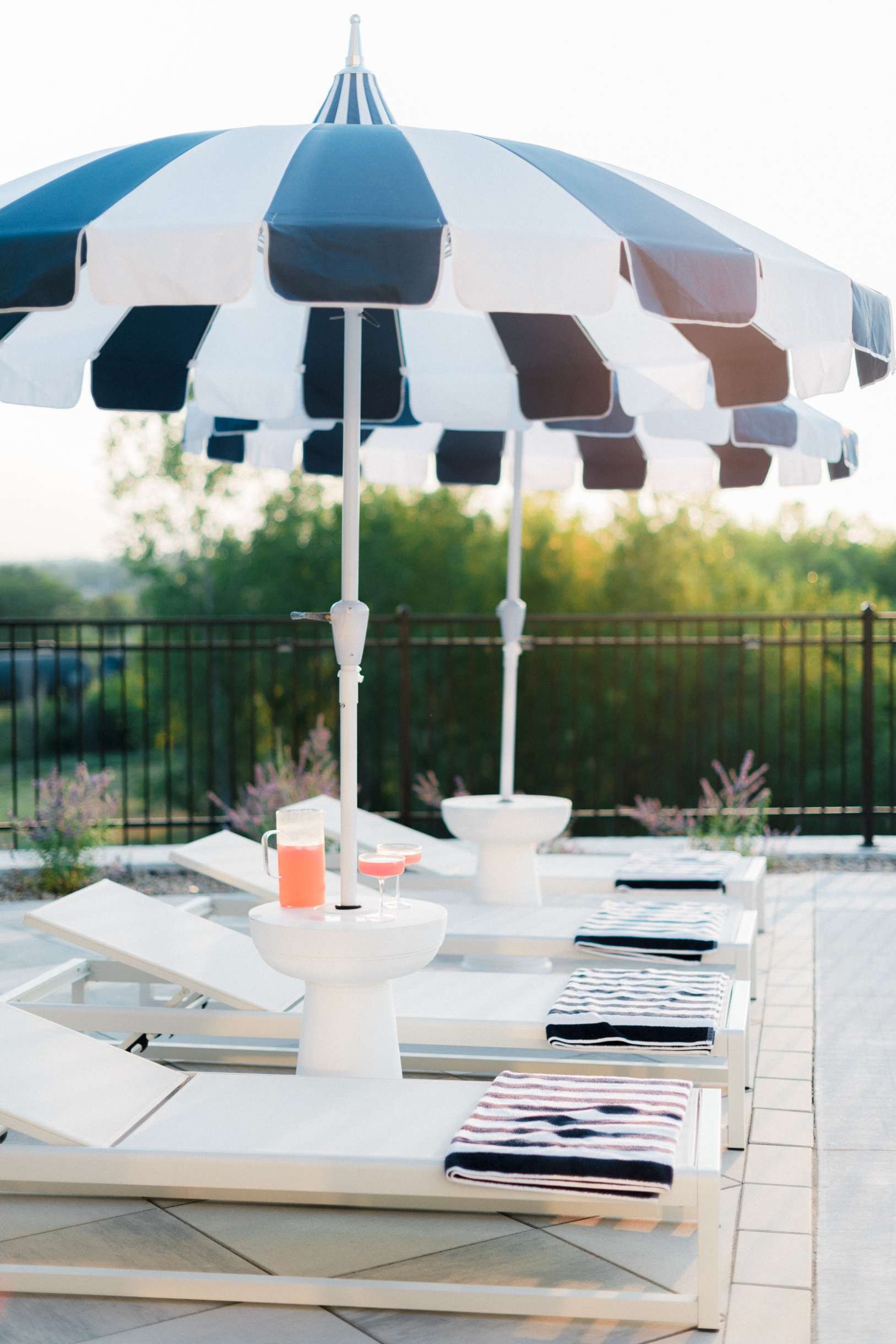 White chaise lounges styled with striped towels and side tables, beneath a large navy and white striped umbrella. Drinks are visible on the side tables