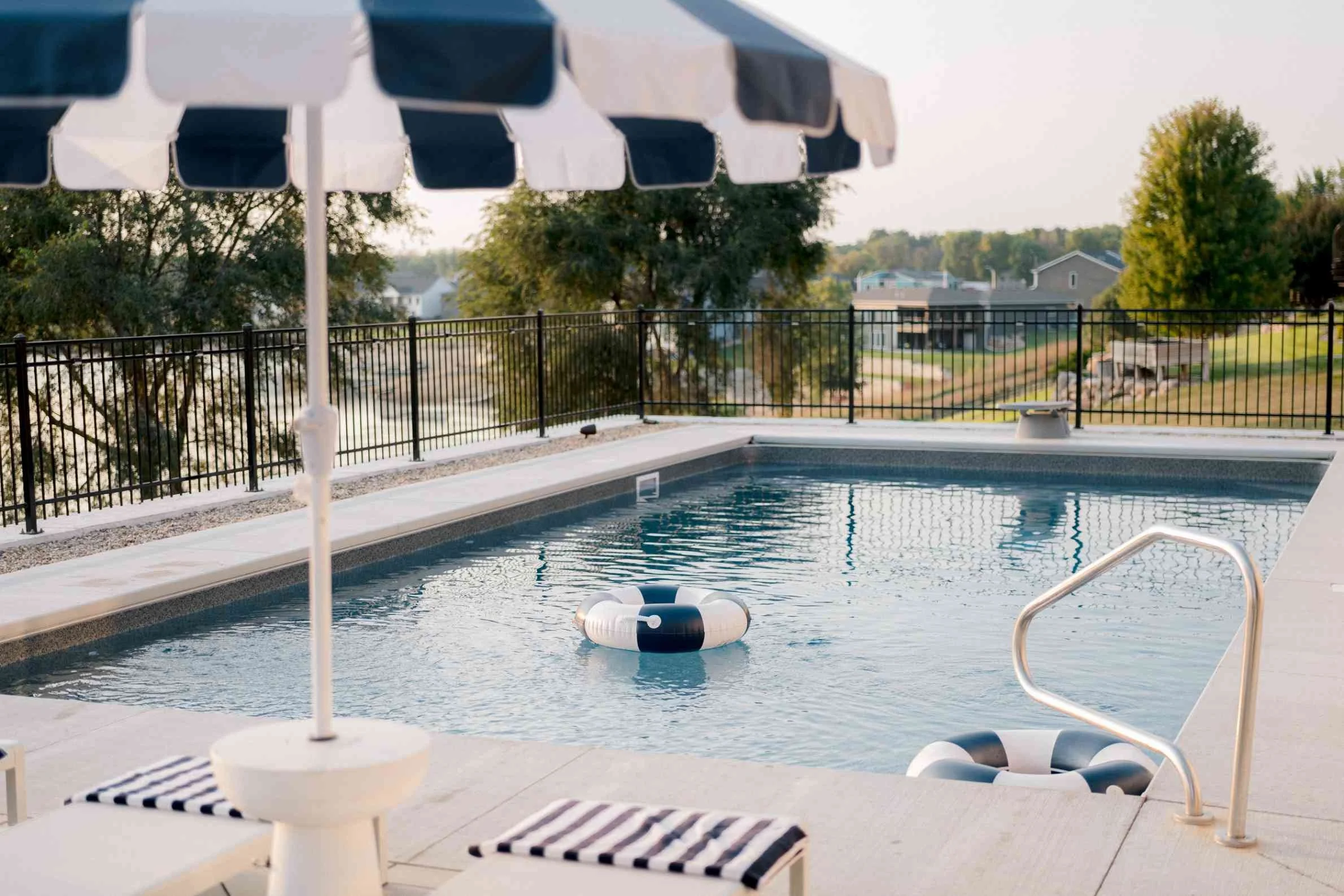 Swimming pool detail showing a white side table, a navy and white striped umbrella, and a ring float in the water. The pool overlooks a body of water in the distance.