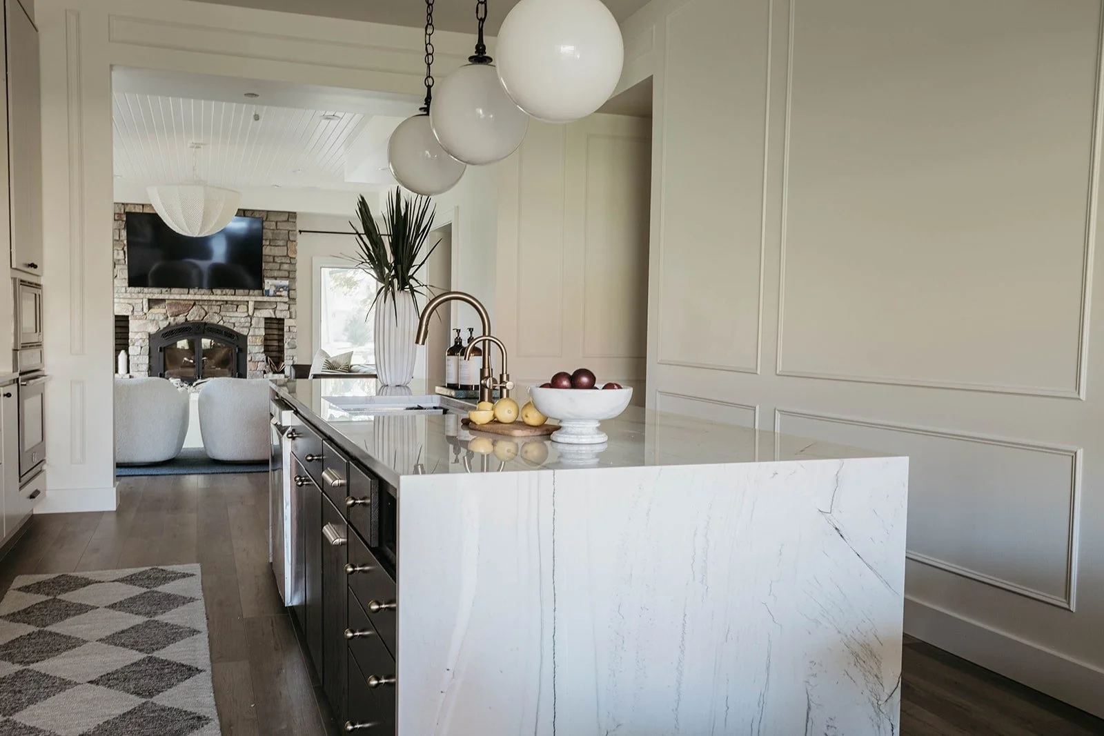 View of the kitchen and living area showing a large white marble waterfall kitchen island and the fireplace in the distance, linking the two spaces.