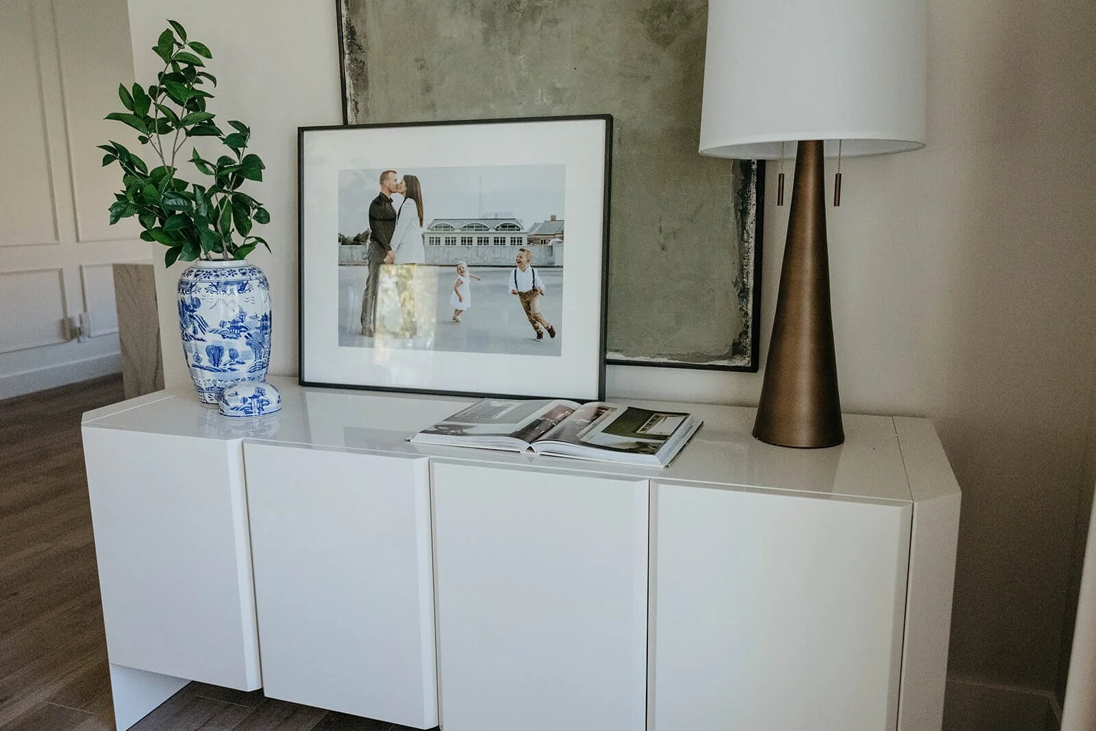 White, paneled credenza styled with framed artwork, a gold table lamp, and a blue and white vase in a living area.