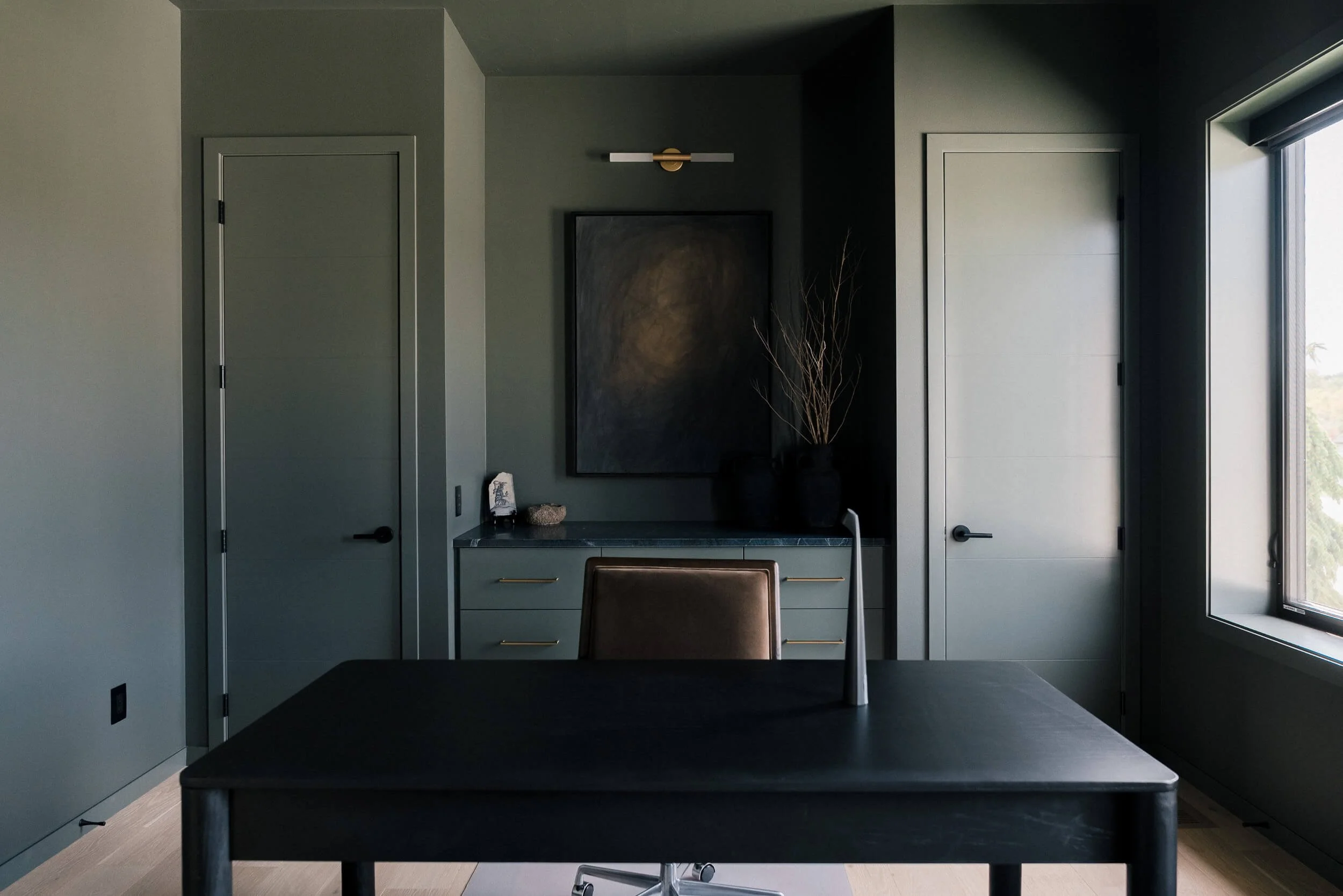 Contemporary home office painted in a deep olive green, featuring a dark wood desk, a brown leather chair, and a dark framed piece of abstract art.
