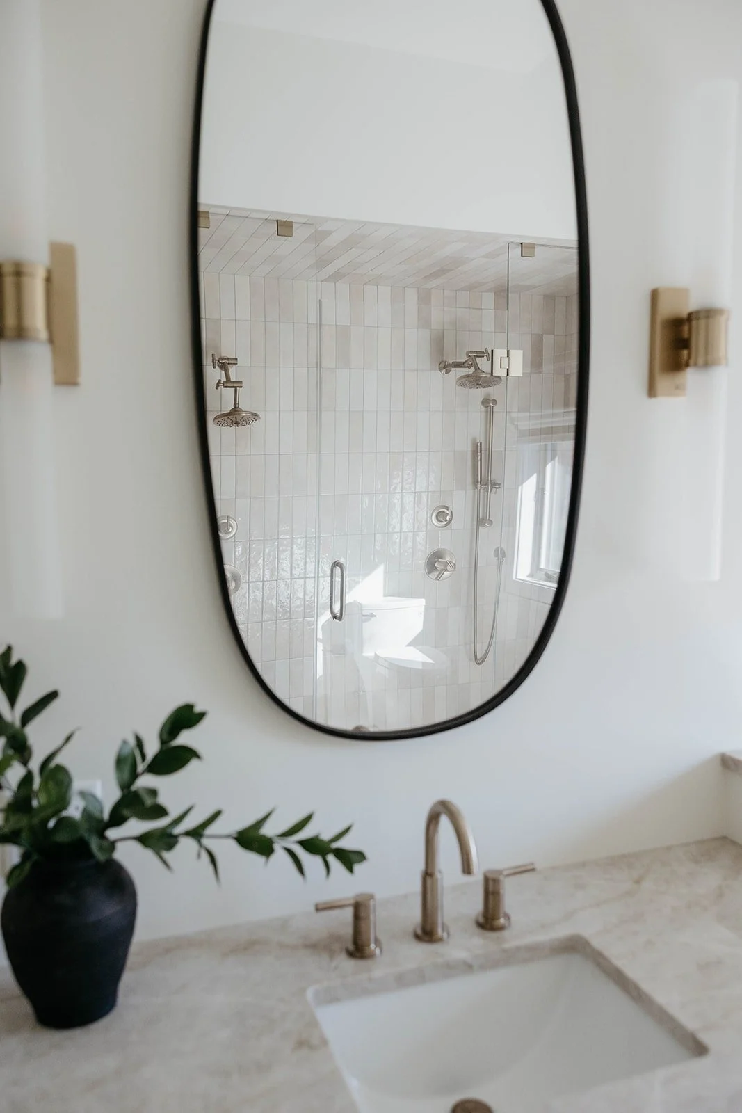 Reflection in the black oval mirror showing the vertical tile and brass shower system in the walk-in shower.