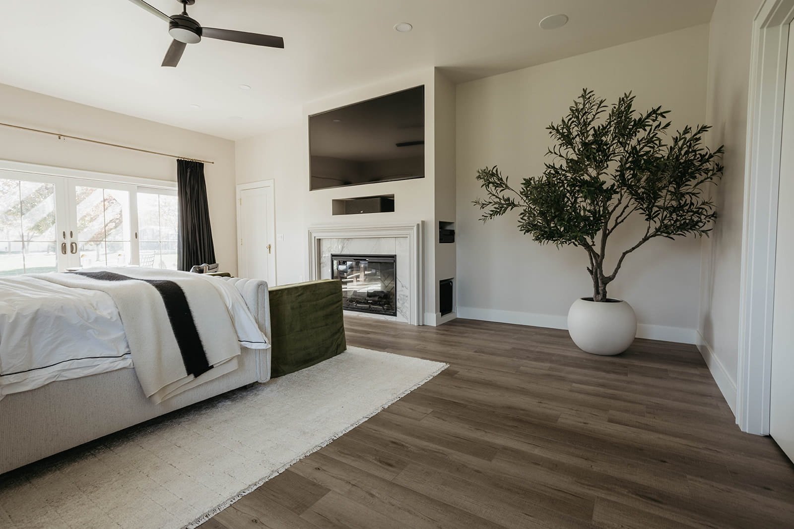 Master bedroom view showing a modern fireplace, a mounted television, a large potted tree, and a dark green accent bench at the foot of the bed.