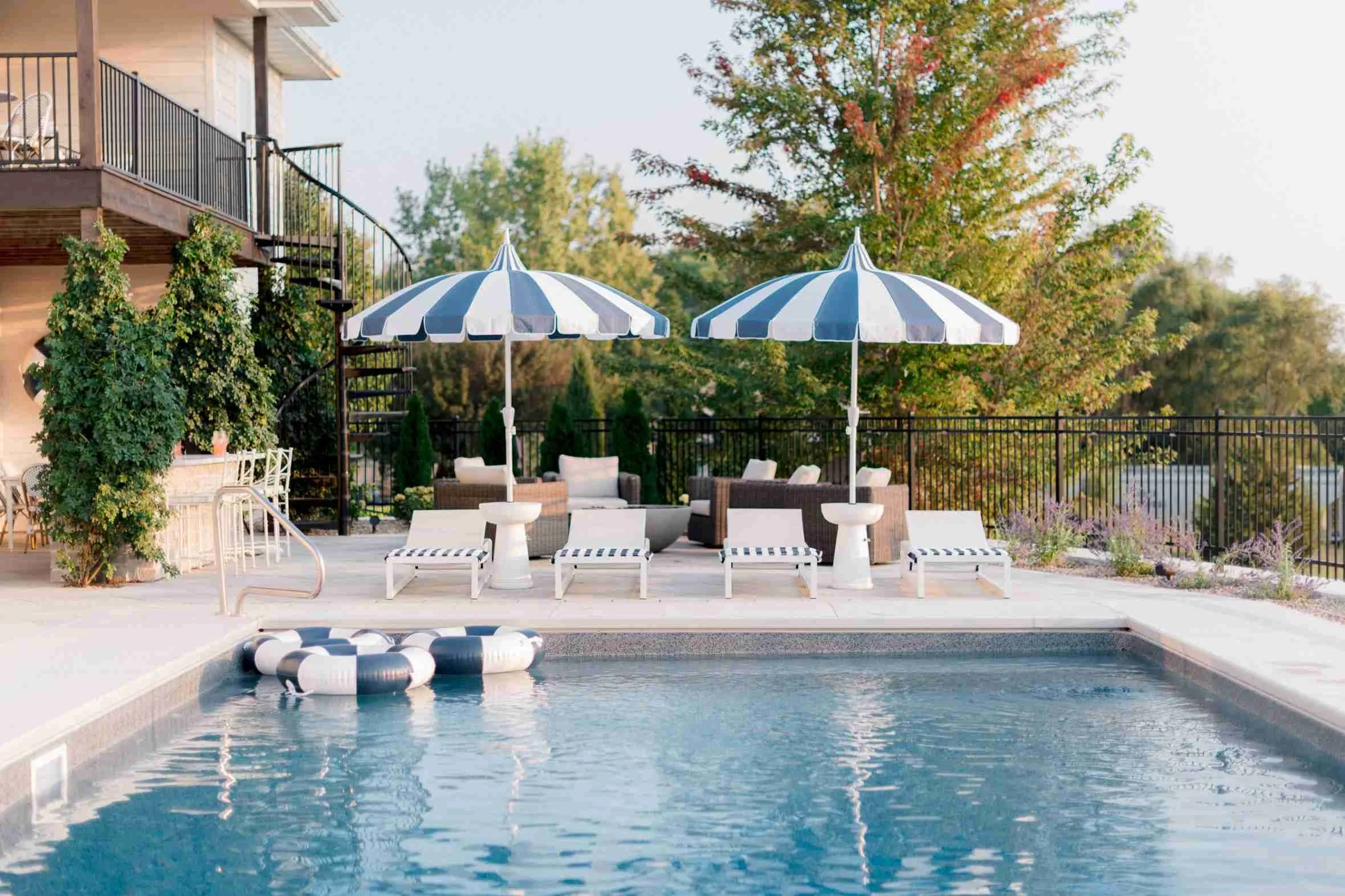 View of the rectangular swimming pool and the back of a multi-story home, showing white lounge chairs, two striped umbrellas, and pool floats.