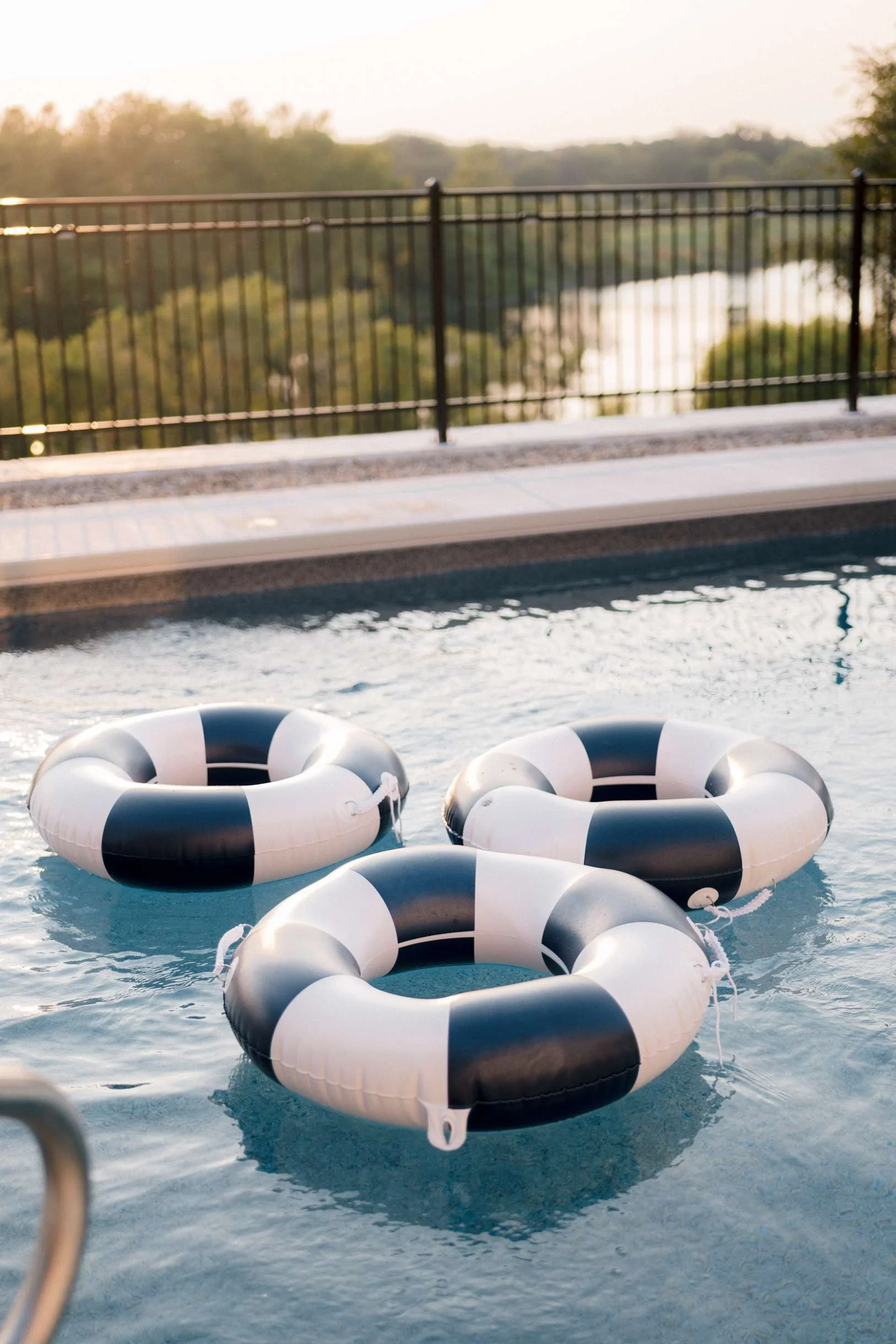 Three navy and white striped pool floats clustered together in the corner of a swimming pool, with a view of the landscape beyond the black metal fence.