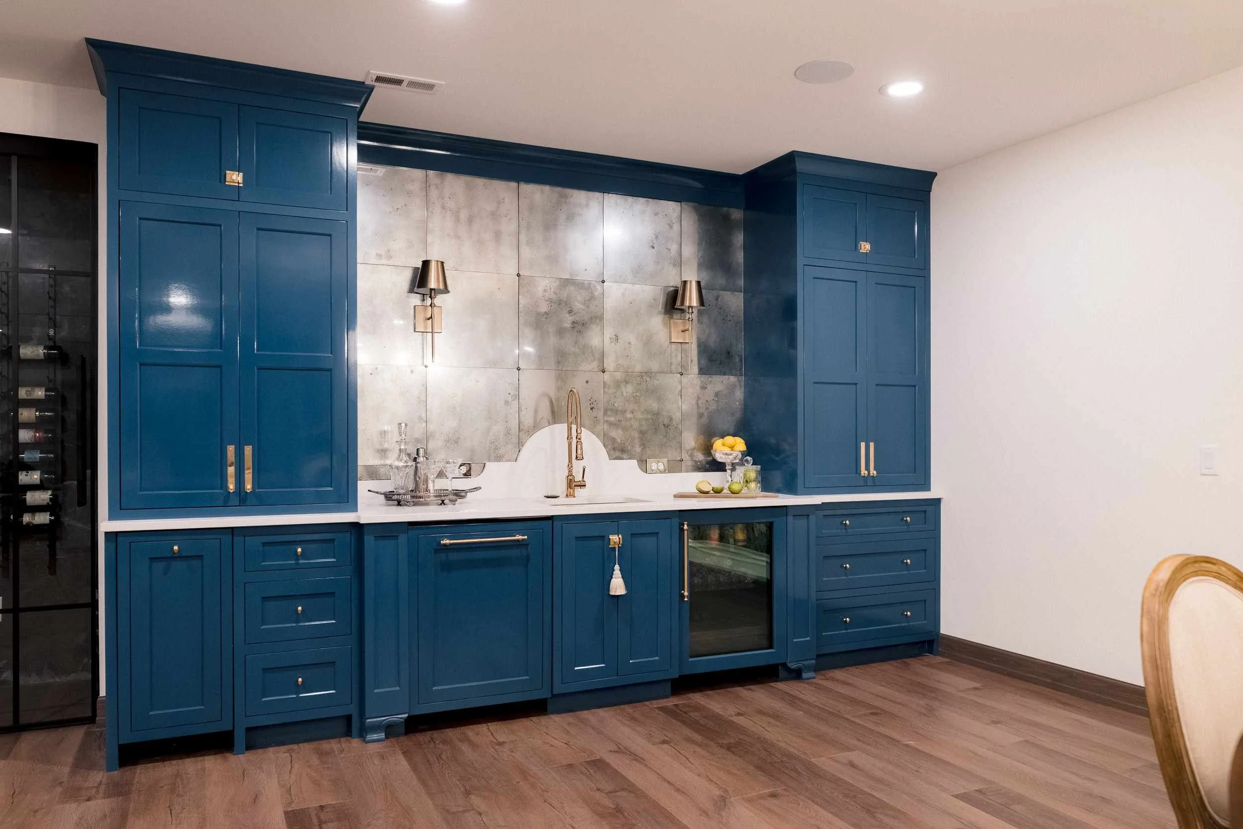 Full view of the blue cabinetry bar, antique metal backsplash, gold sconces, and refrigerator drawers beneath a white counter.