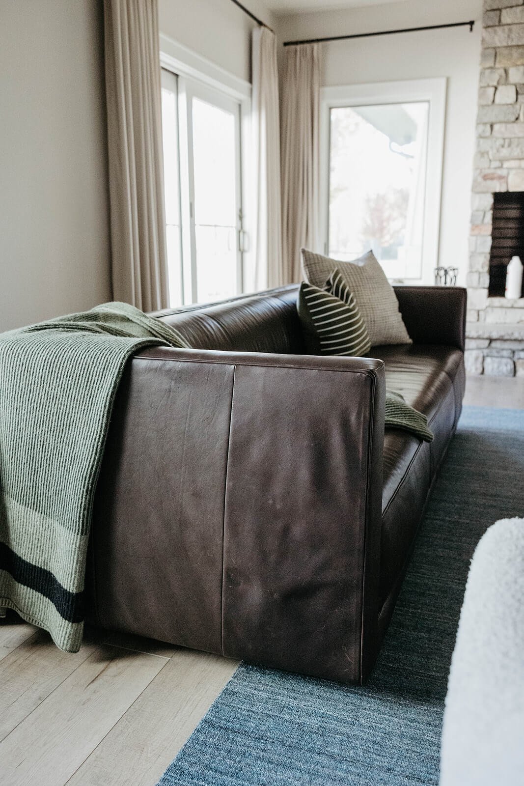 Close-up of a square-armed dark brown leather sofa draped with a soft green knit throw blanket, set on a blue patterned rug near a large window.