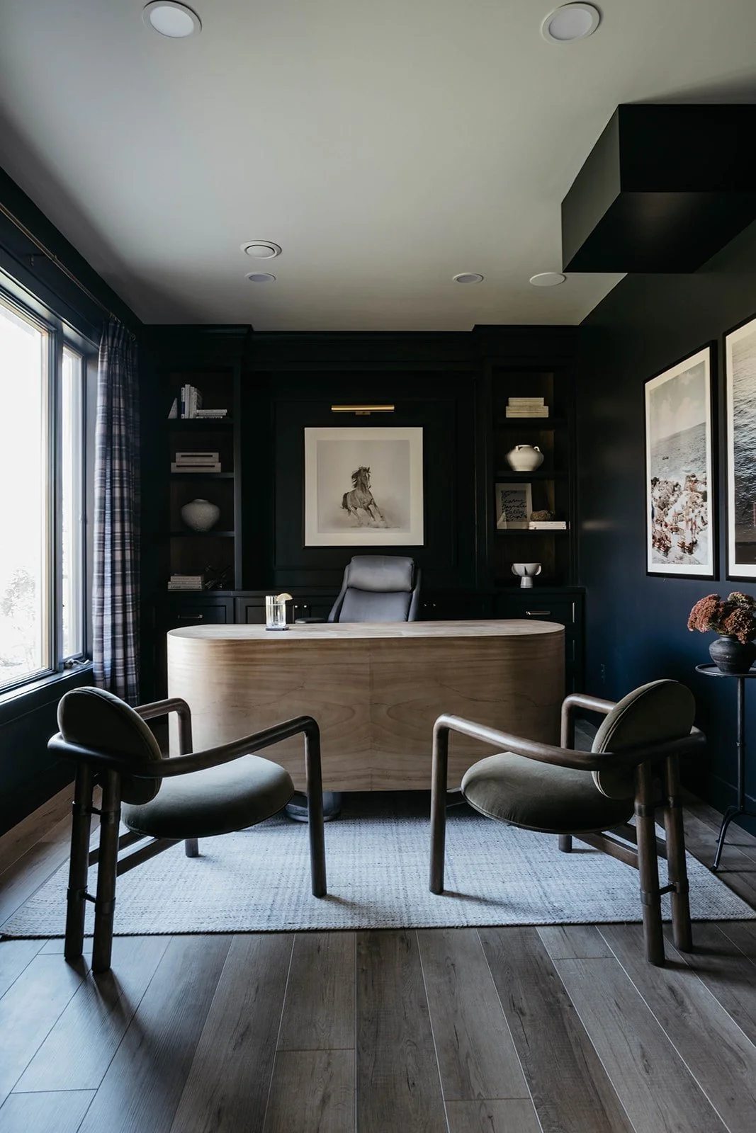 Masculine home office design with dark walls, light wood flooring, a curved light wood desk, and two dark, sculptural chairs on a patterned rug.
