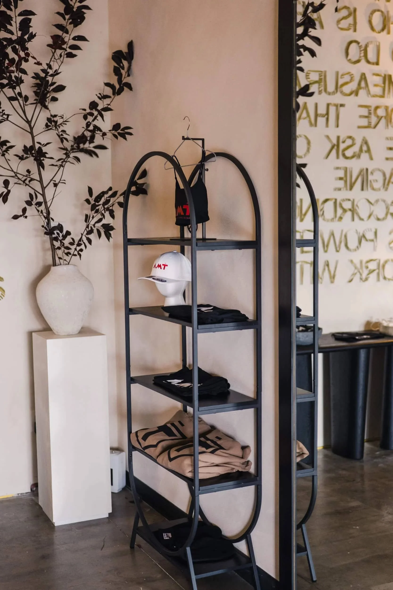 Full view of the black arched metal retail display rack with hats and small bags, with dark foliage in a white vase on a white pedestal sitting next to it.