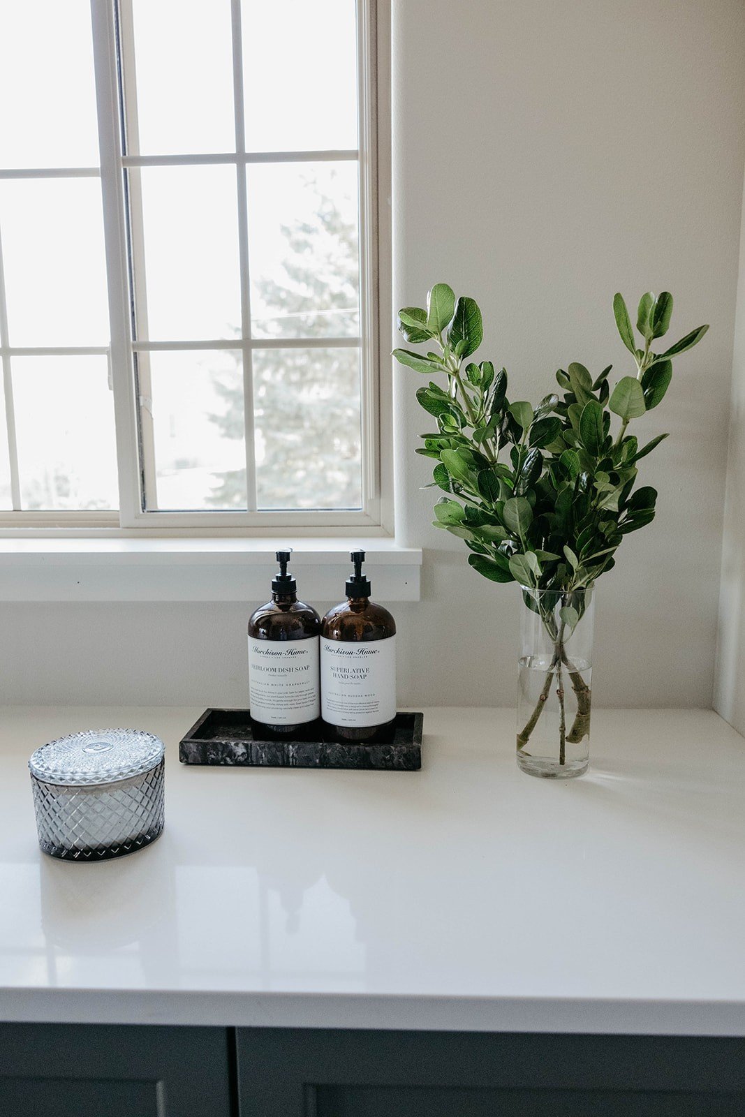 Close-up of the laundry room window and the white countertop styled with soap bottles on a black tray and a vase of fresh greenery.